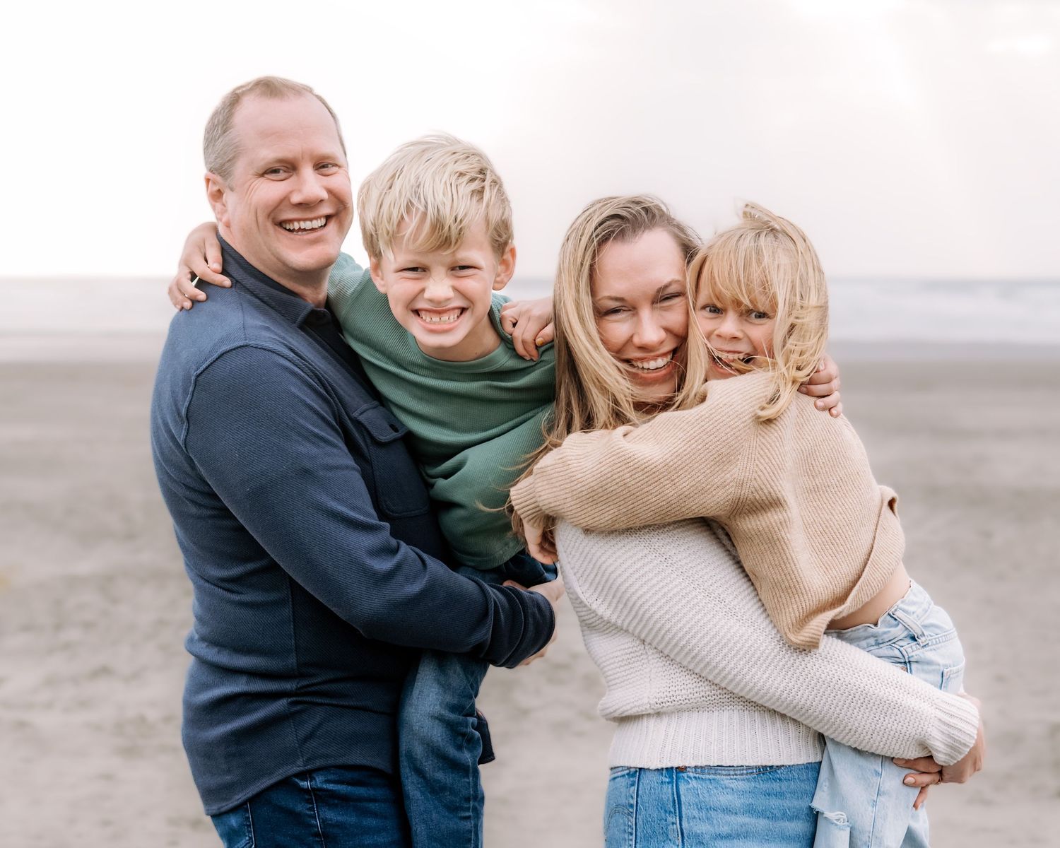 Happy family of four smiling and hugging on a beach, parents carrying two young blonde children outdoors.