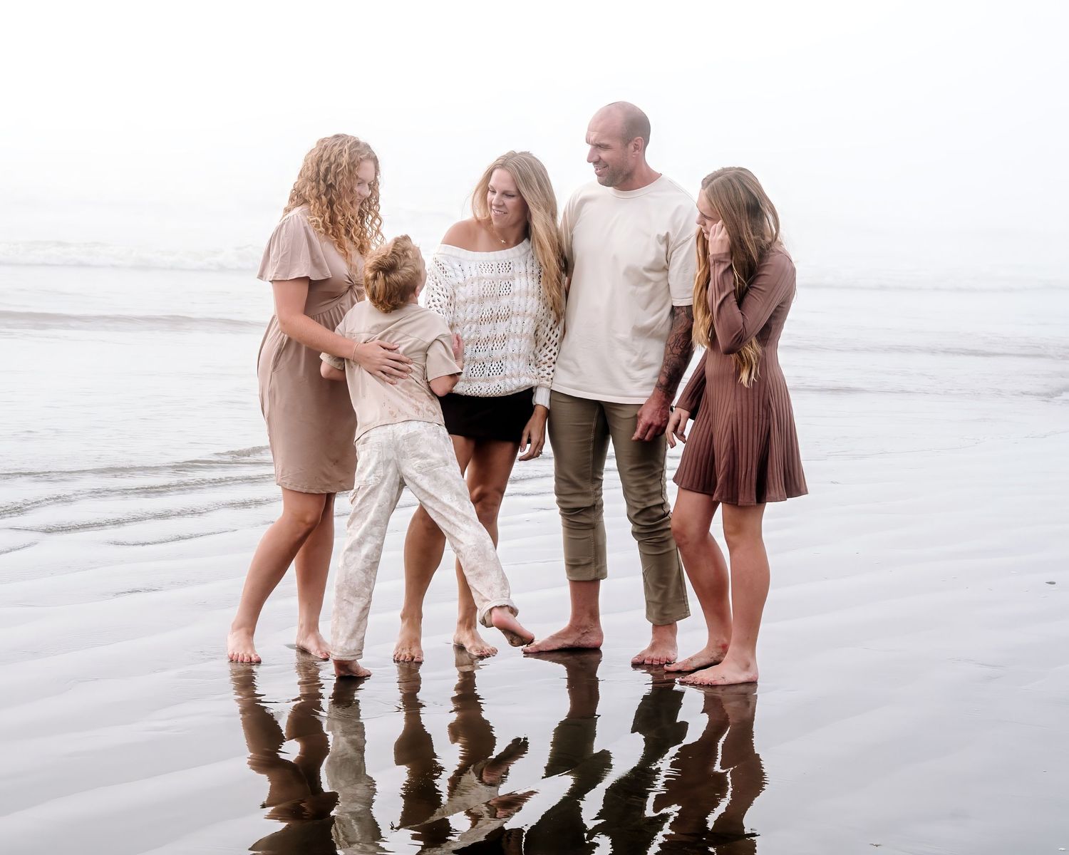 Family of five in casual neutral outfits stand barefoot together on a wet sandy beach with misty ocean waves and reflections behind them.
