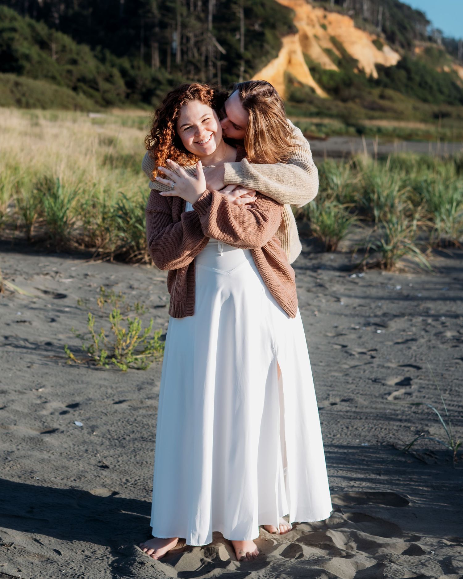 Two people share a warm embrace on a beach with coastal cliffs in background.