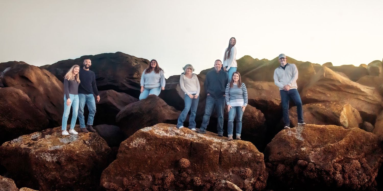Group of friends in casual clothing standing together on large rust-colored coastal rocks at sunset.