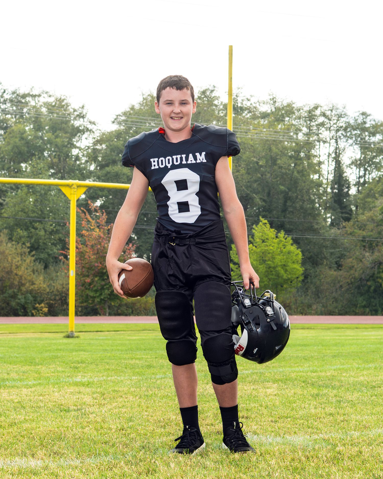 High school football player in black jersey number 8 poses with ball and helmet on field.