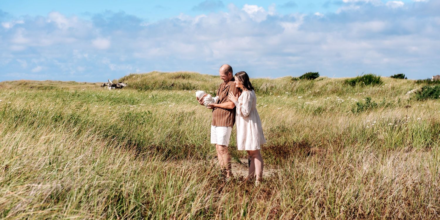 Couple standing together in tall golden grass on a windswept coastal dune under a blue sky with scattered white clouds.