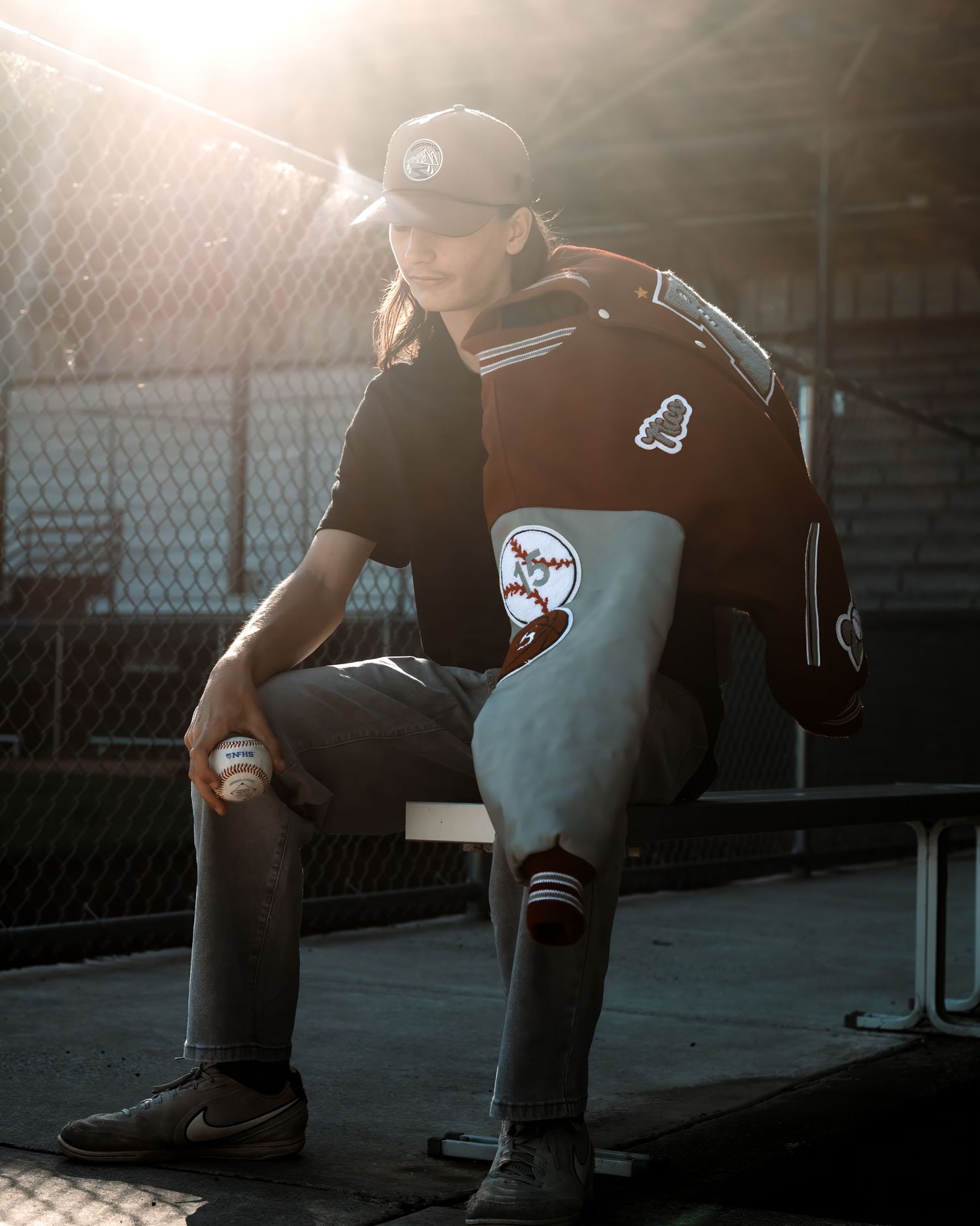 Young baseball player in a cap and maroon letterman jacket sits on a dugout bench holding a baseball as sunlight streams through the fence.