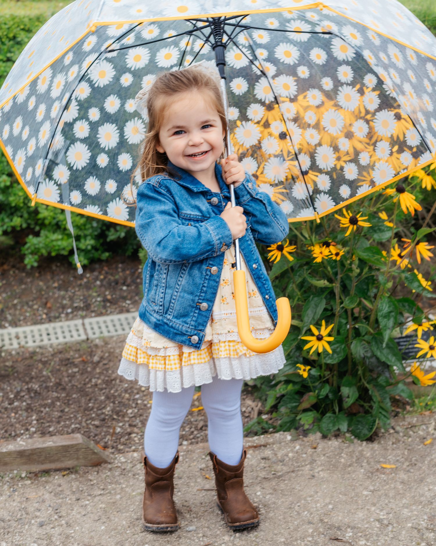 Child holding polka dot umbrella poses near yellow flowers in garden setting.