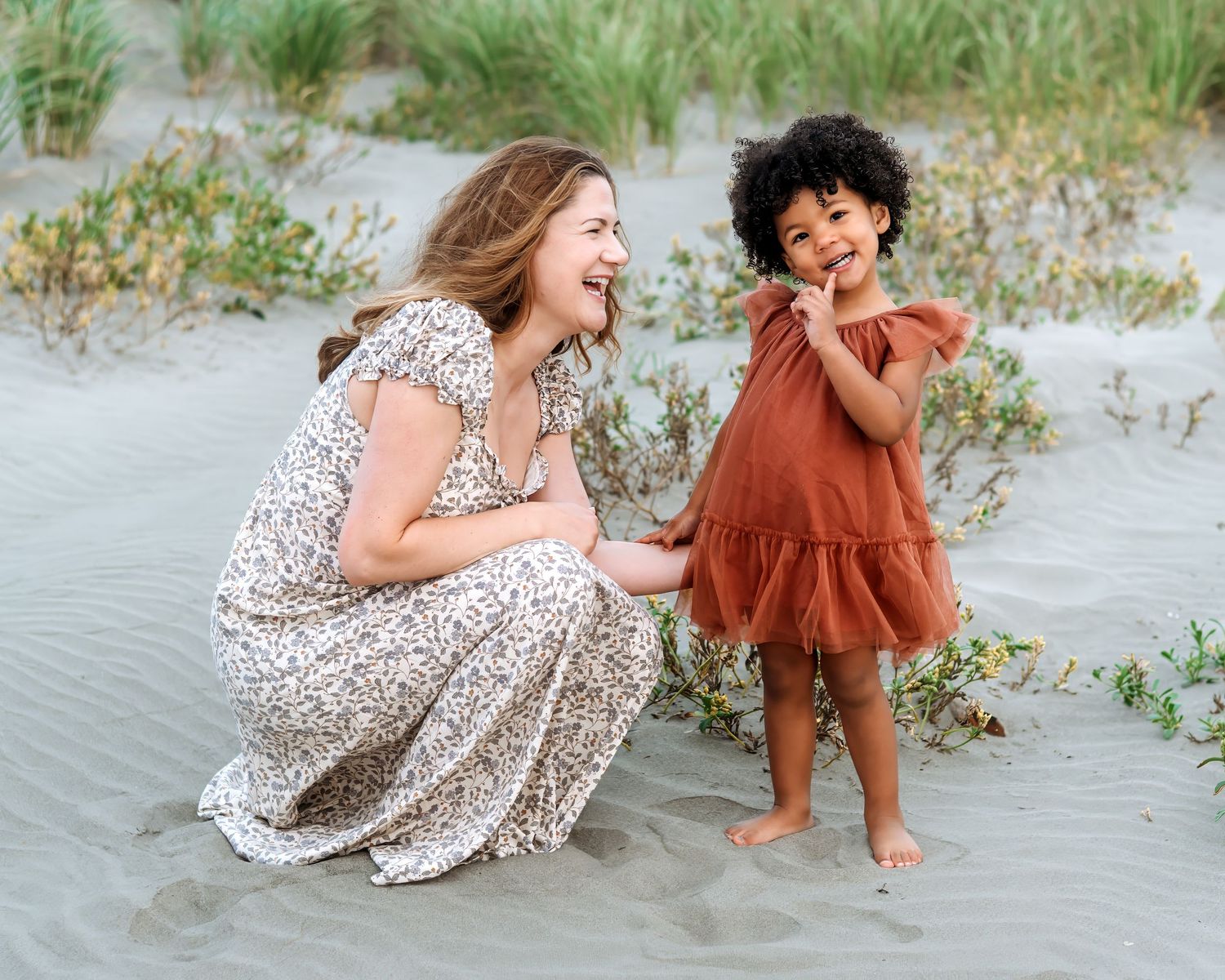 A woman in a floral dress crouches smiling beside a young girl in a rust dress on sandy beach dunes.
