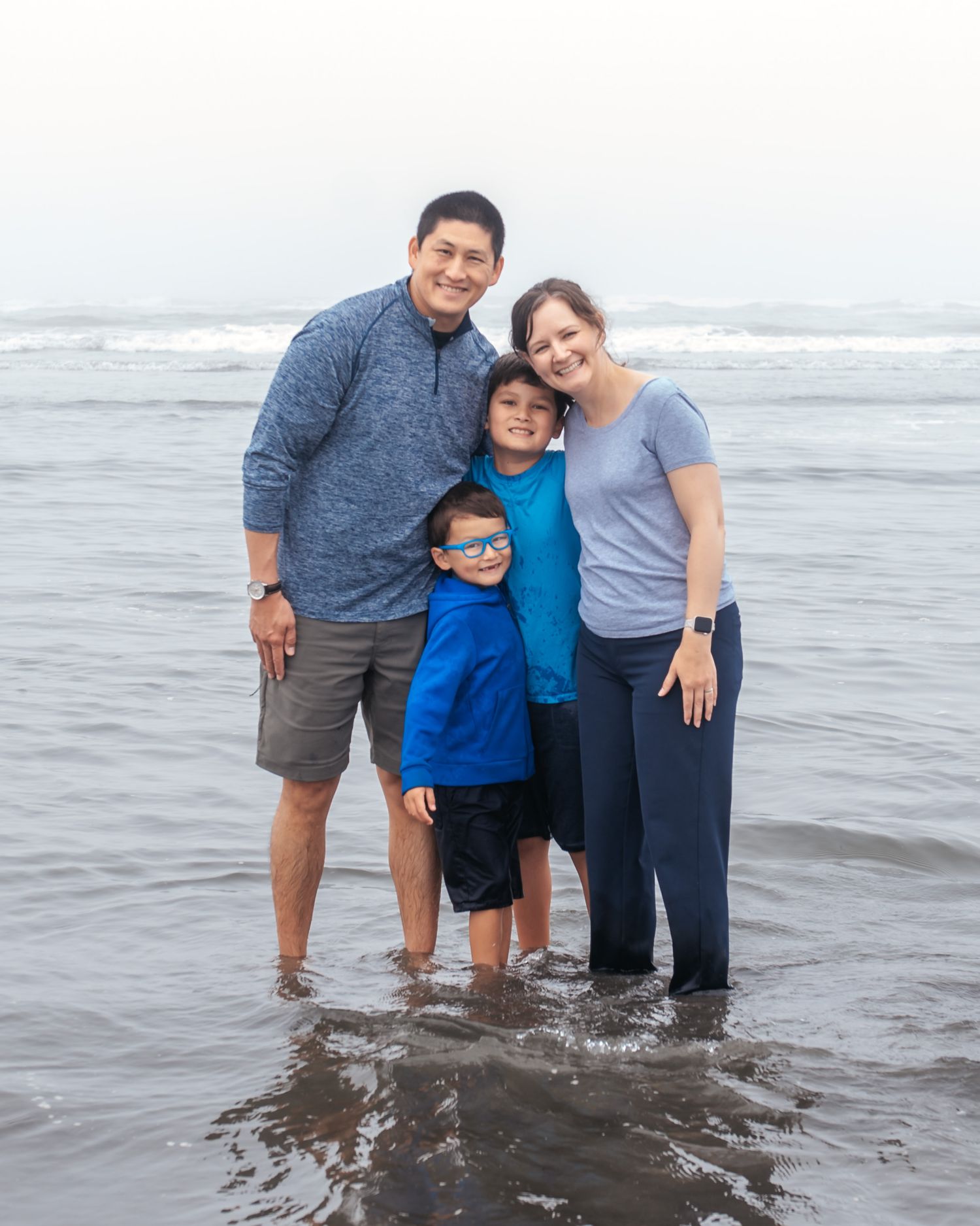 A family of four stands together at the beach with waves and ocean in the background.
