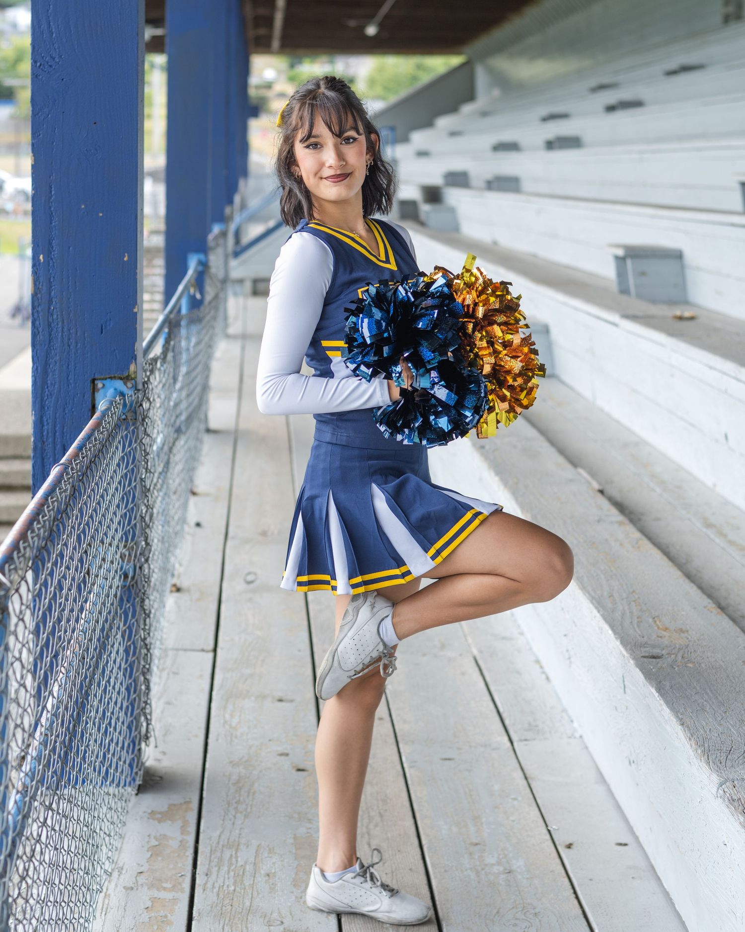 A cheerleader performs poses in a blue and gold uniform under stadium bleachers.