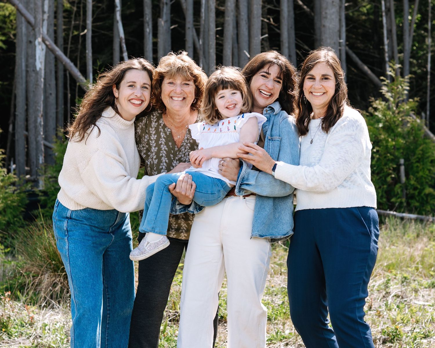 Five women smile outdoors near pine trees, with one holding a young child in a joyful family portrait.