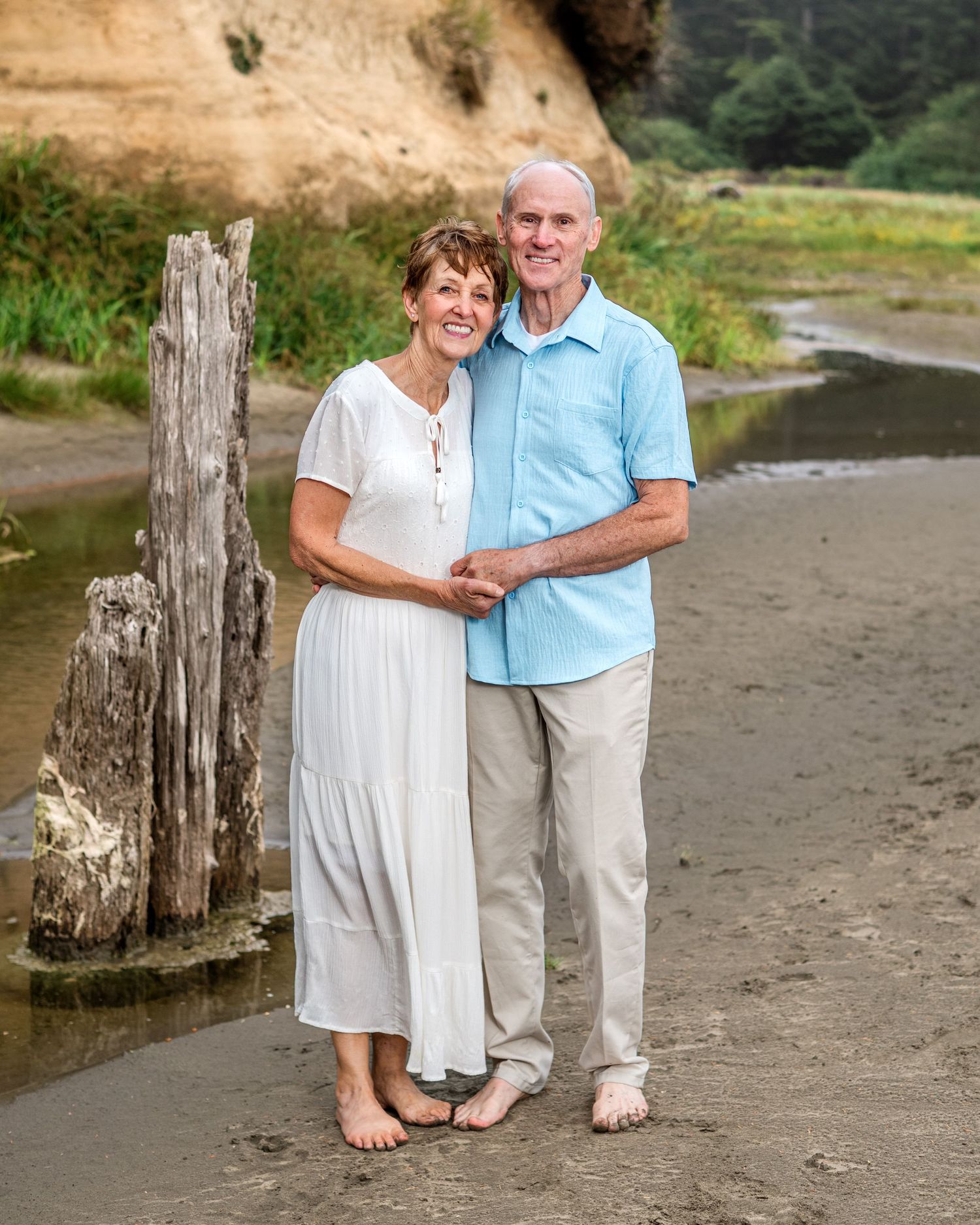 Couple poses barefoot on sandy beach with wooden posts.