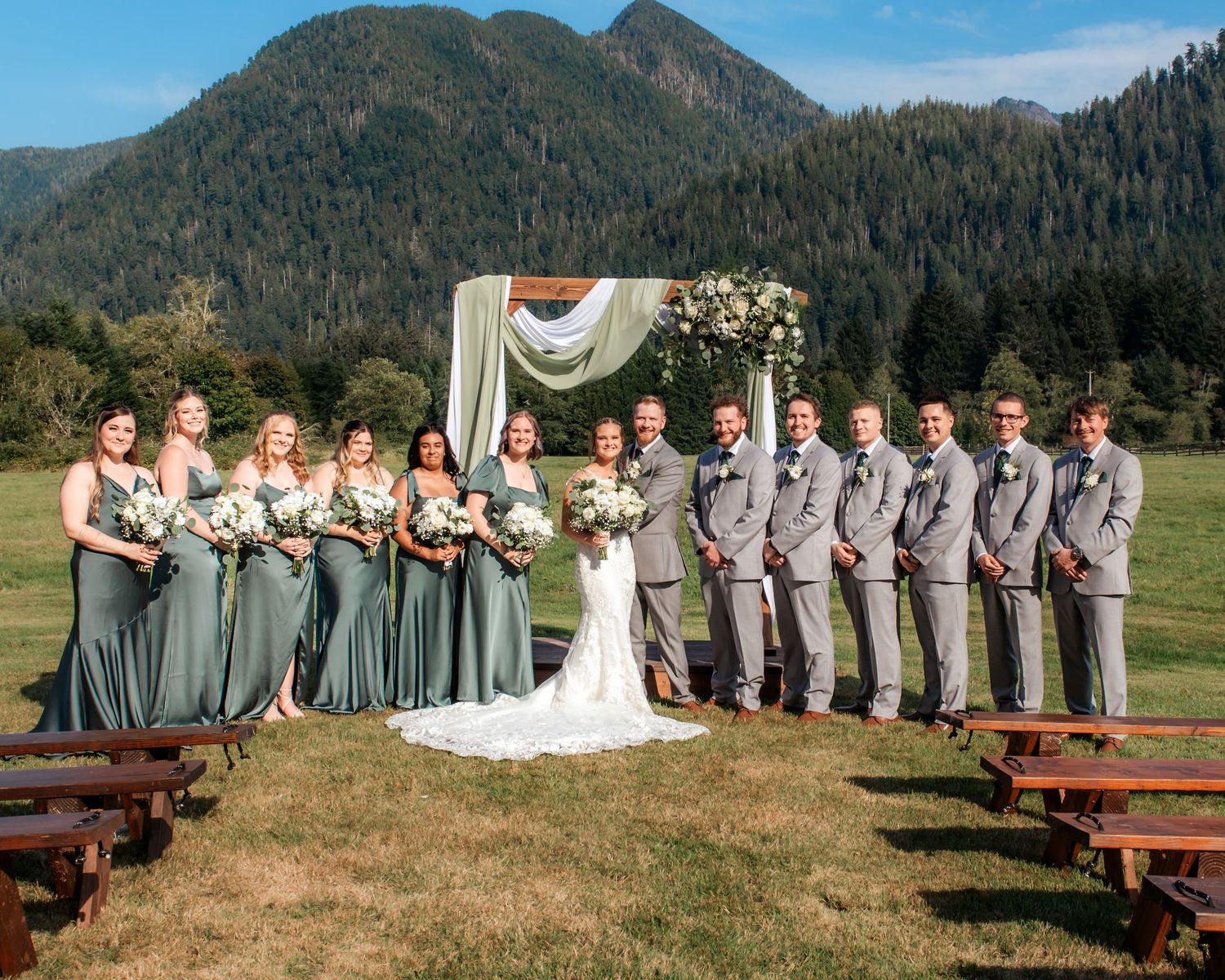 Wedding party lined up outdoors with mountain landscape and wooden benches.