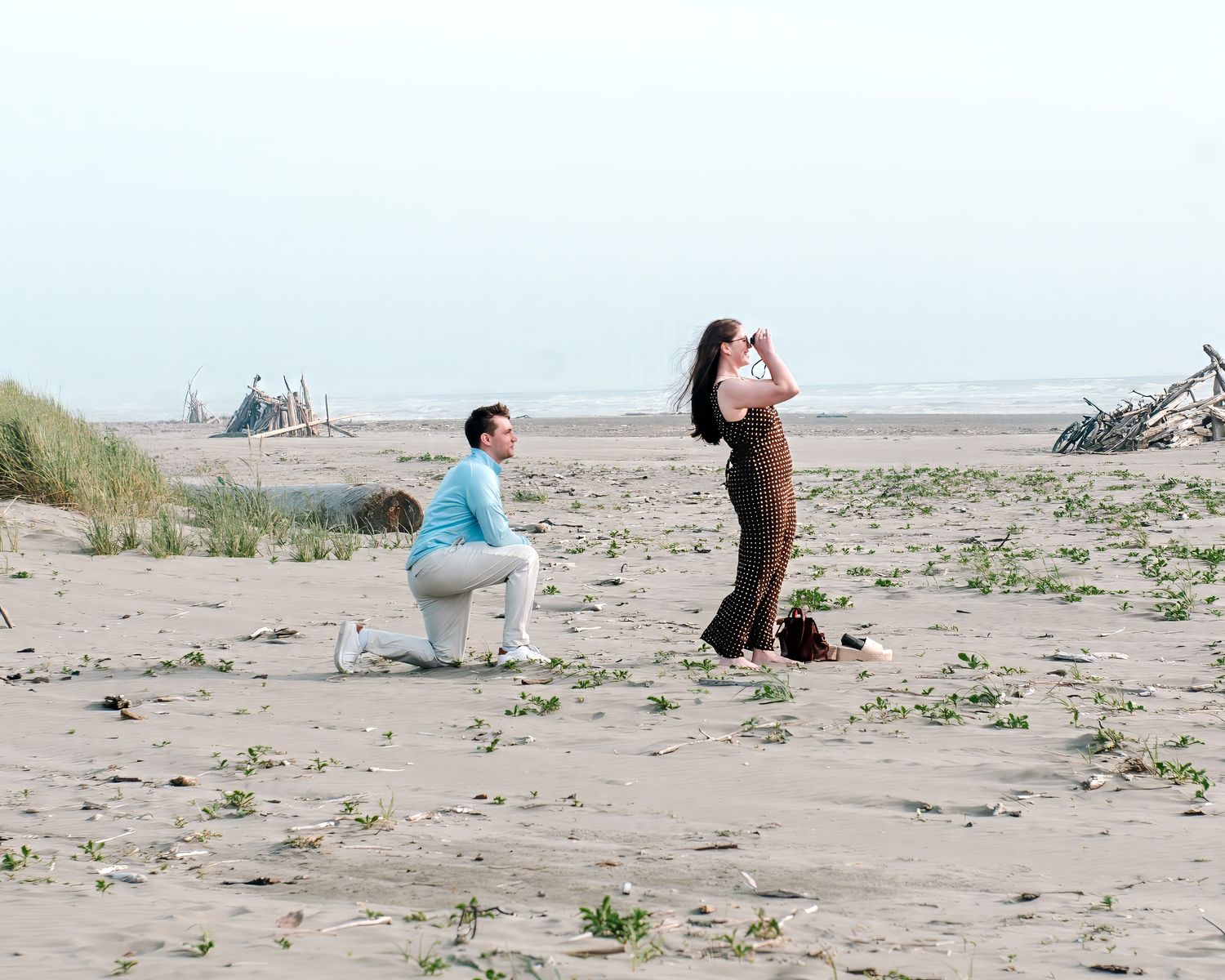 Man kneeling on sandy beach proposing to woman looking through binoculars near ocean shore.