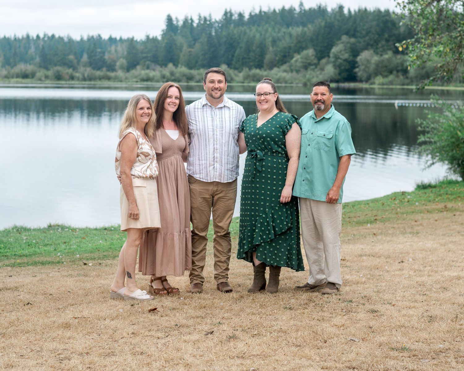 Five people smiling together outdoors near a serene lake surrounded by trees on a cloudy day.