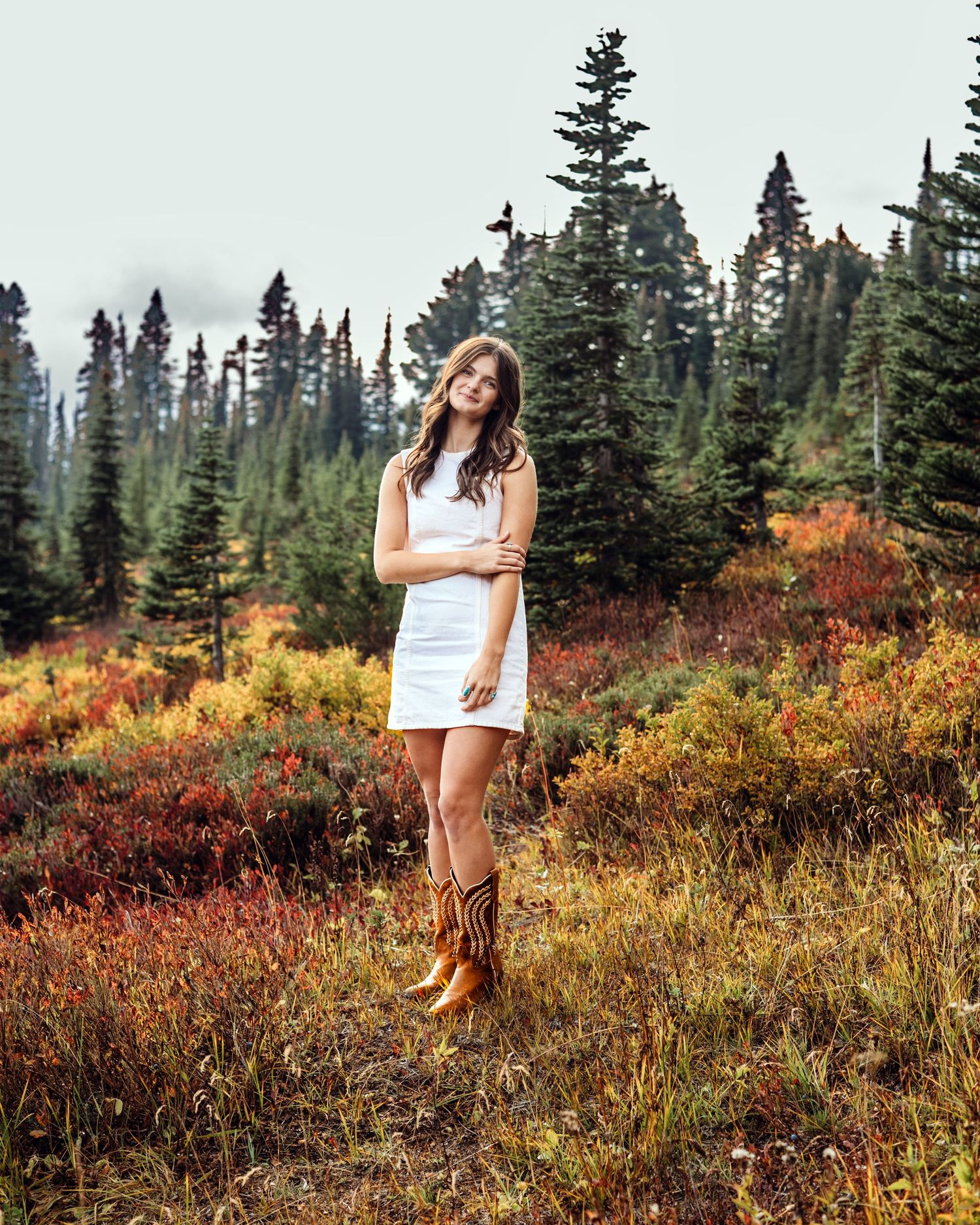 Person in white dress and brown boots poses in scenic mountain meadow with evergreen trees and autumn foliage.