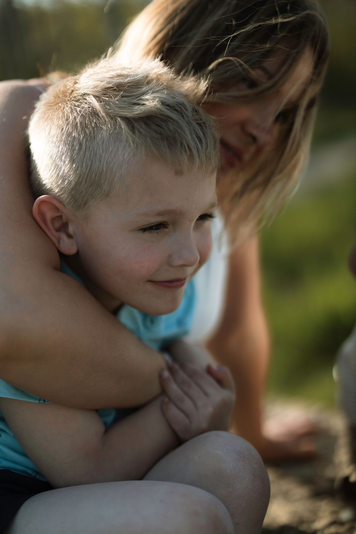 A woman tenderly hugs a young blonde boy outdoors in warm sunlight, sharing a gentle moment of affection.