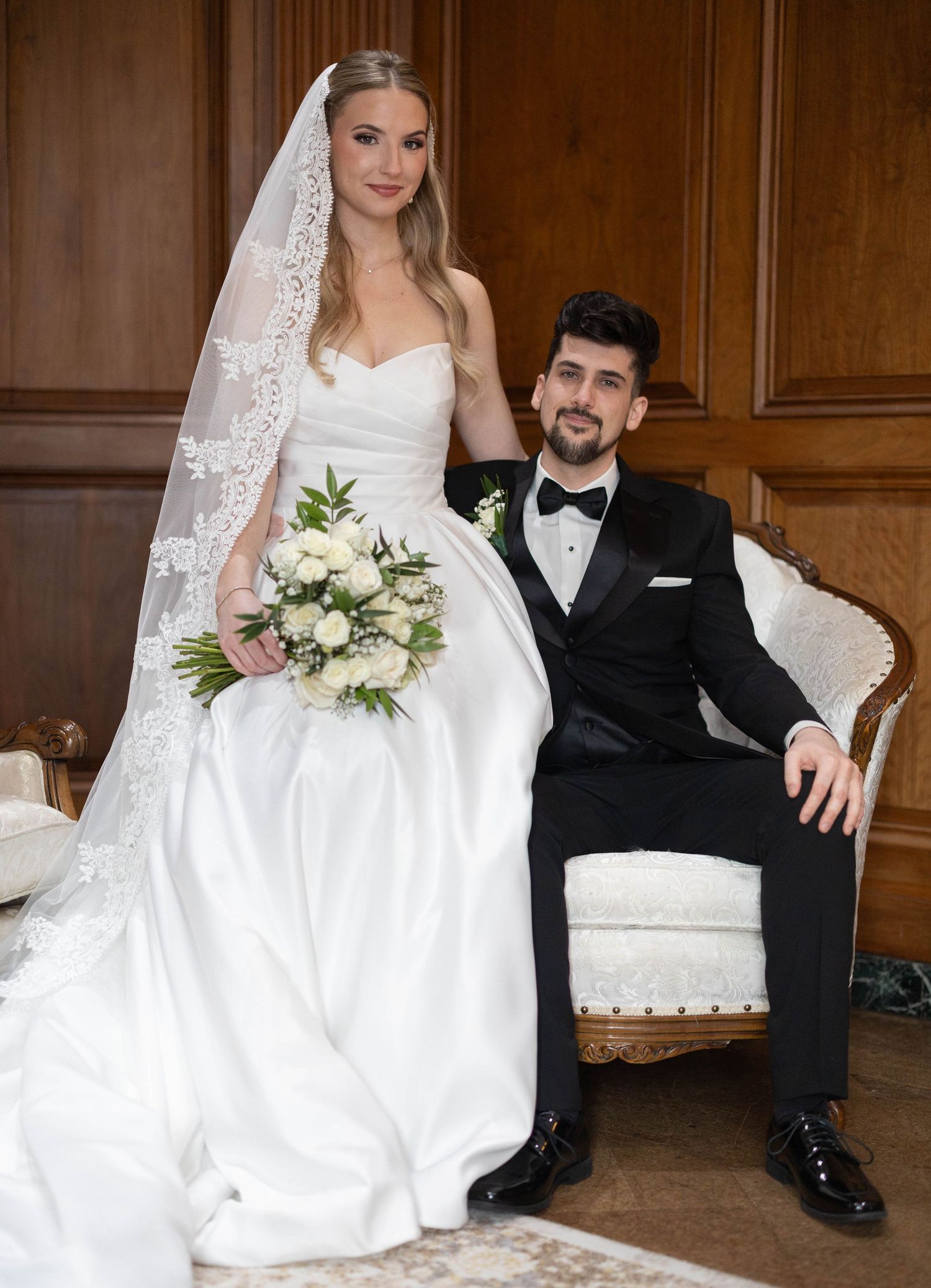 Elegant bride in white gown with lace veil holds bouquet beside groom in black tuxedo at formal wedding portrait.