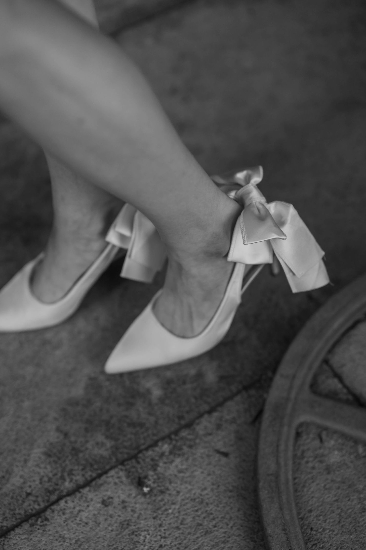 Black and white photo of elegant bridal heels with satin bow detail on a woman's feet.