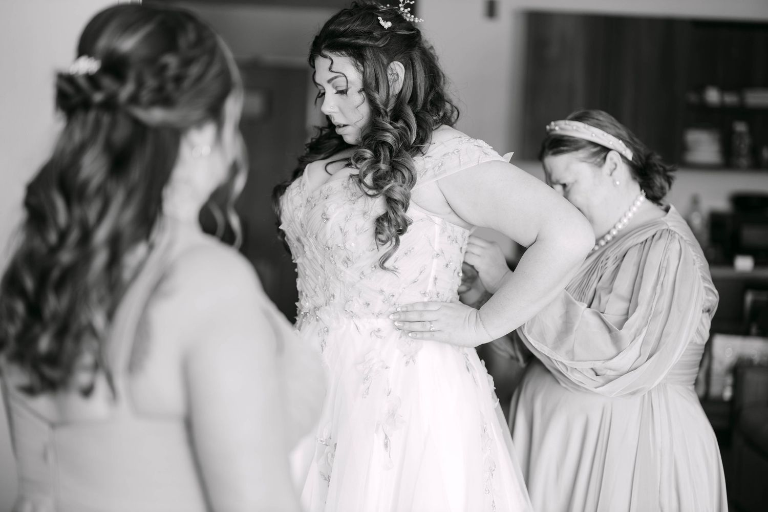 Black and white photo of bride being helped into wedding gown by bridesmaids before ceremony.