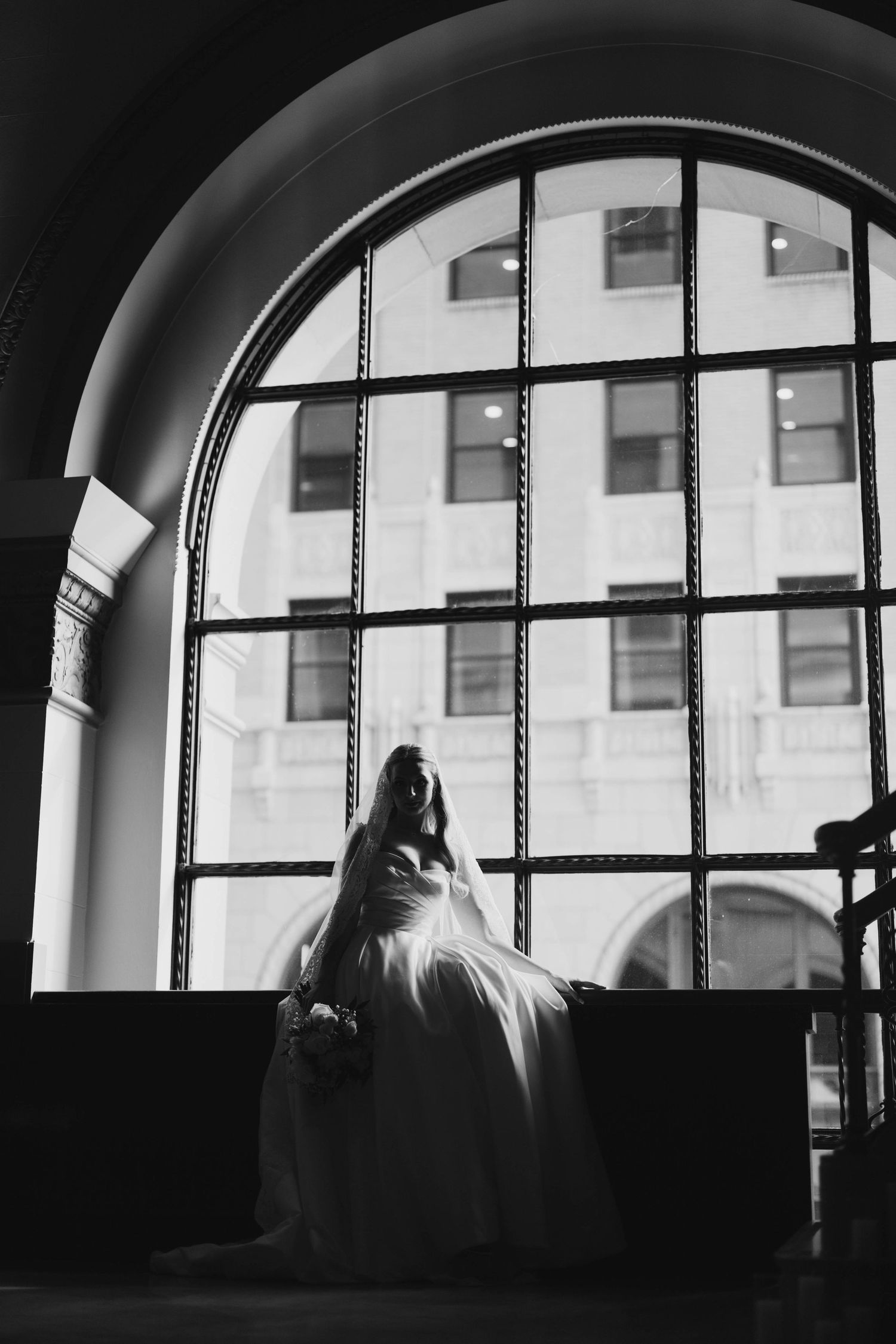 Black and white silhouette of a bride in a wedding gown standing before a large arched window.