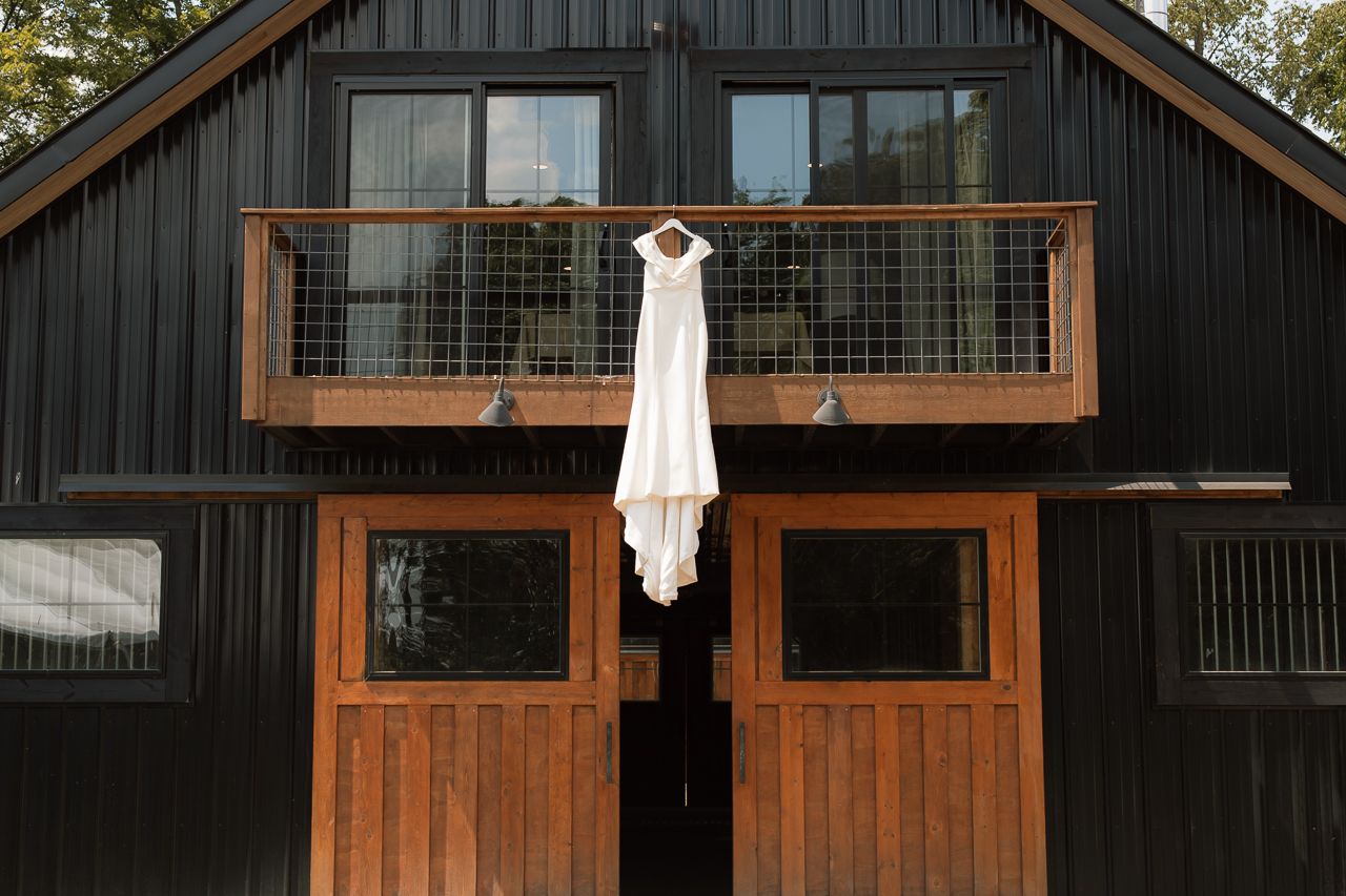 White wedding dress hanging on a hanger from a rustic dark barn balcony with wooden doors.