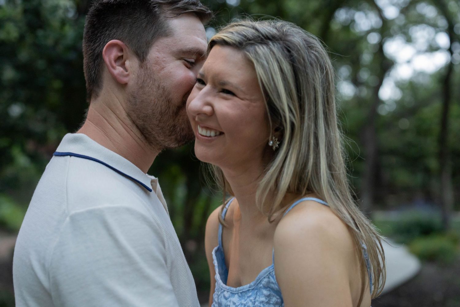 A man tenderly kisses a smiling blonde woman on the cheek outdoors among lush green trees.