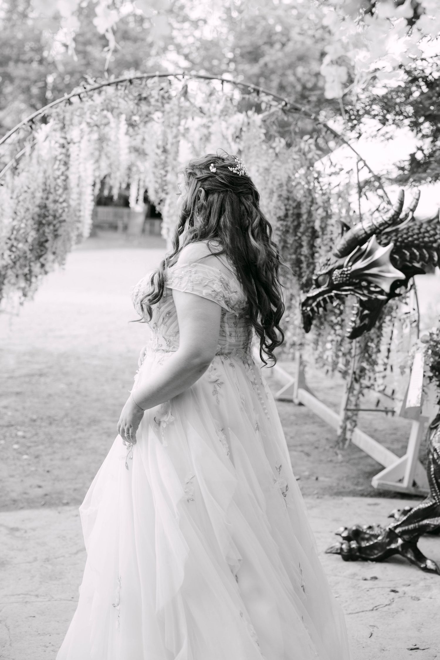 Black and white photo of a bride in a flowing wedding gown standing under a floral arch with a dragon sculpture nearby.
