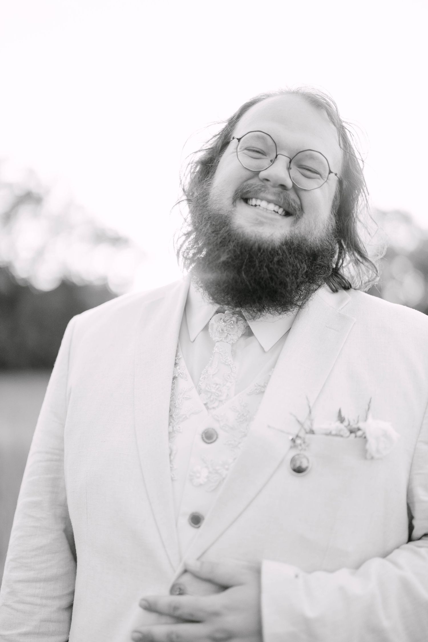 Black and white portrait of a bearded groom in a white suit, smiling joyfully with round glasses and a boutonniere.