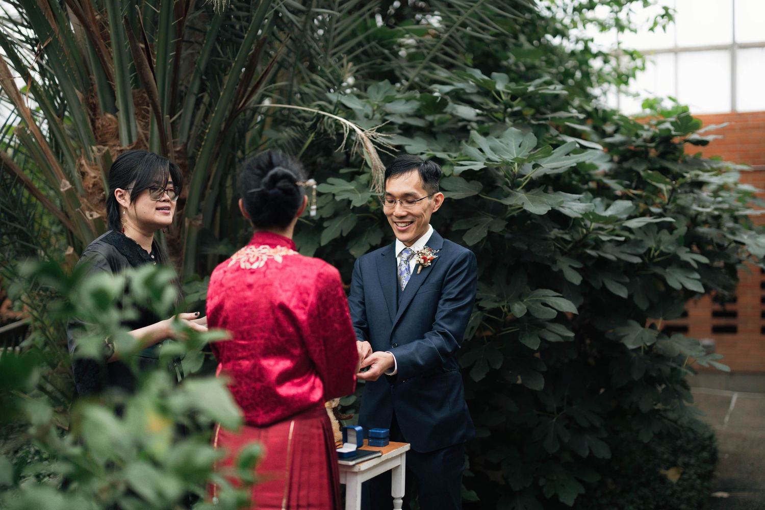 Couple in traditional Chinese wedding attire exchanging vows in a lush indoor botanical garden greenhouse.