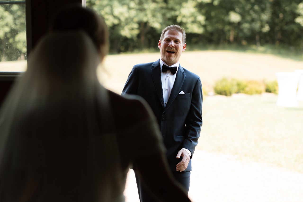 Groom in a classic black tuxedo and bowtie reacts with joy during a first look moment at an outdoor wedding.