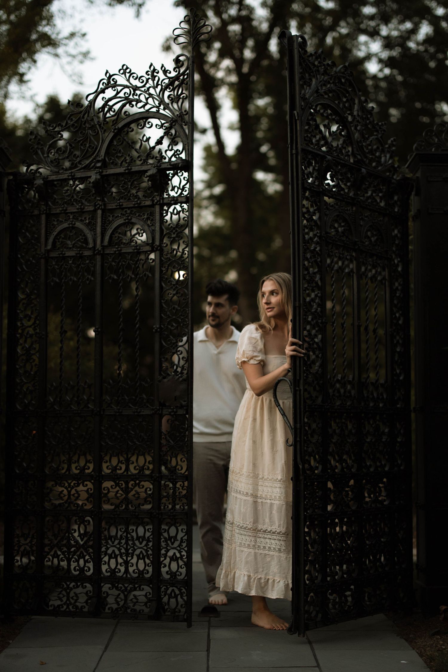 An ornate black iron gate stands in a garden setting during twilight hours.