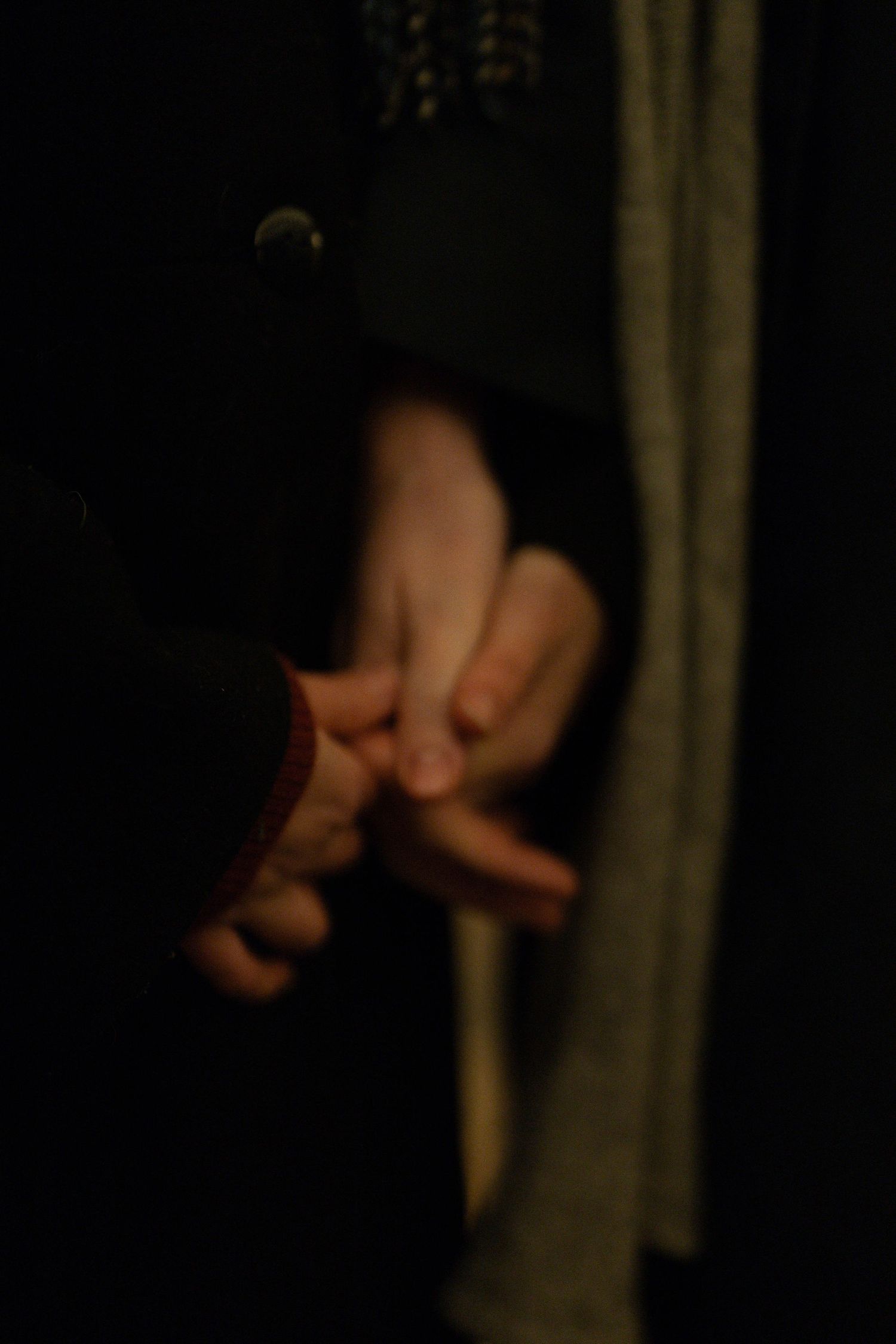 Two people holding hands gently in a dimly lit setting, wearing dark coats and a light scarf.