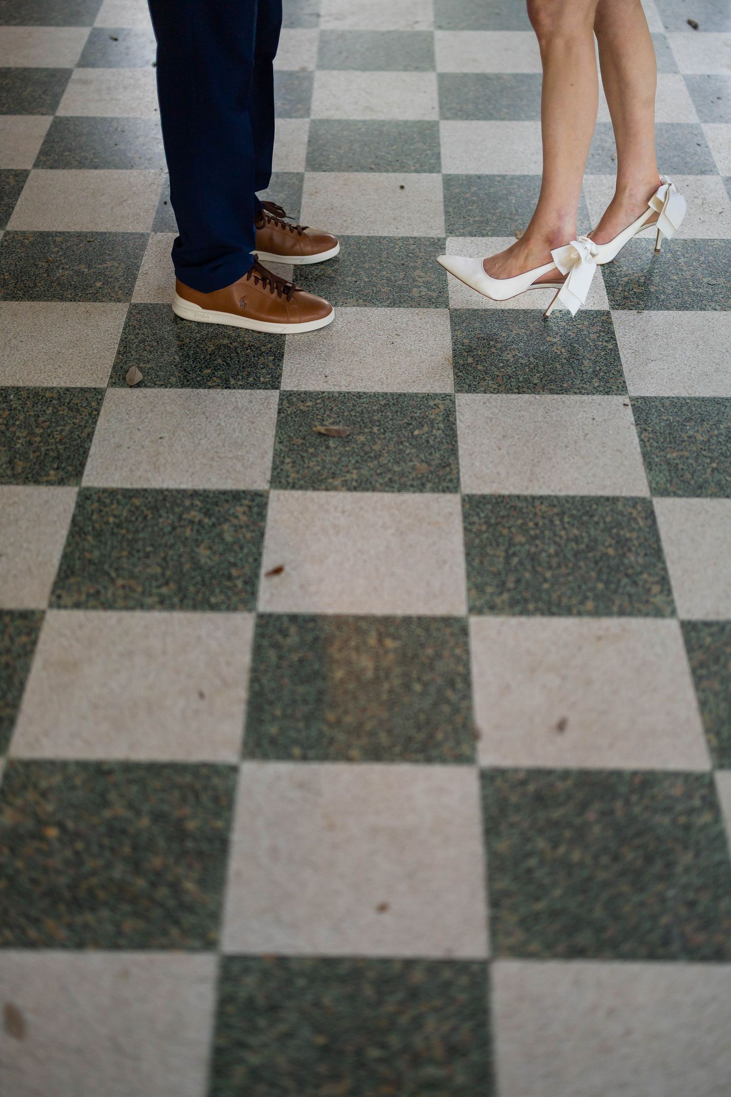Close-up of couple's feet on checkered floor, man in brown sneakers and woman in white bow heels.