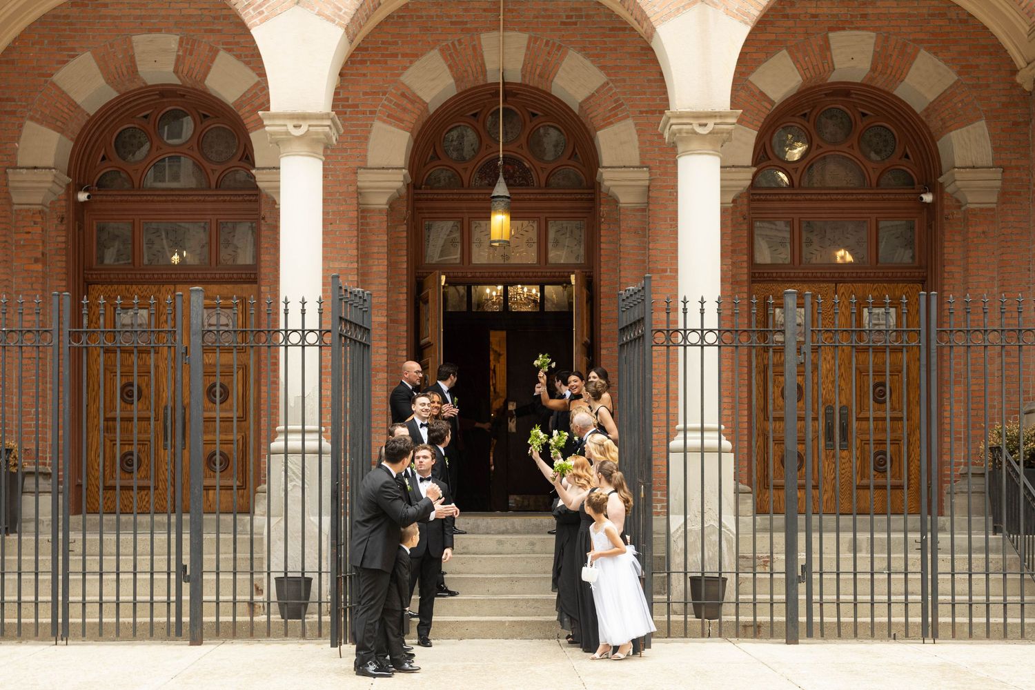 Wedding party celebrating outside elegant brick church with arched doorways and iron fence after ceremony.
