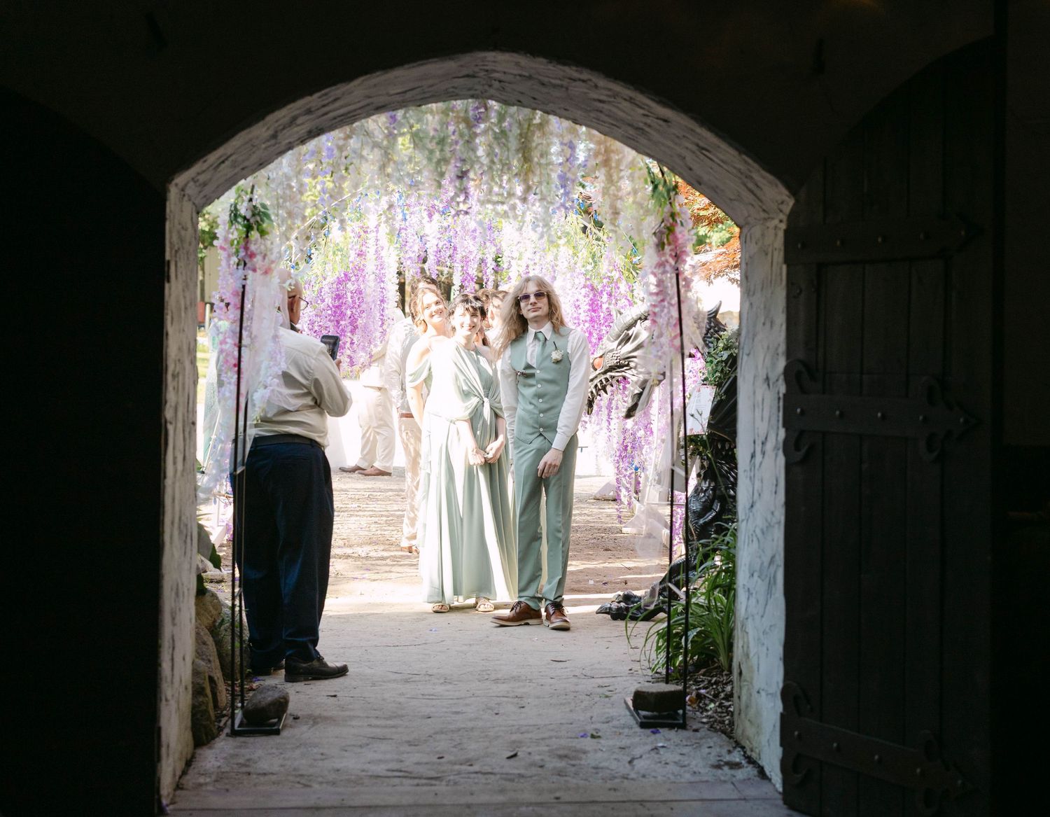 People standing under a floral arch with purple wisteria blooms in a garden courtyard.