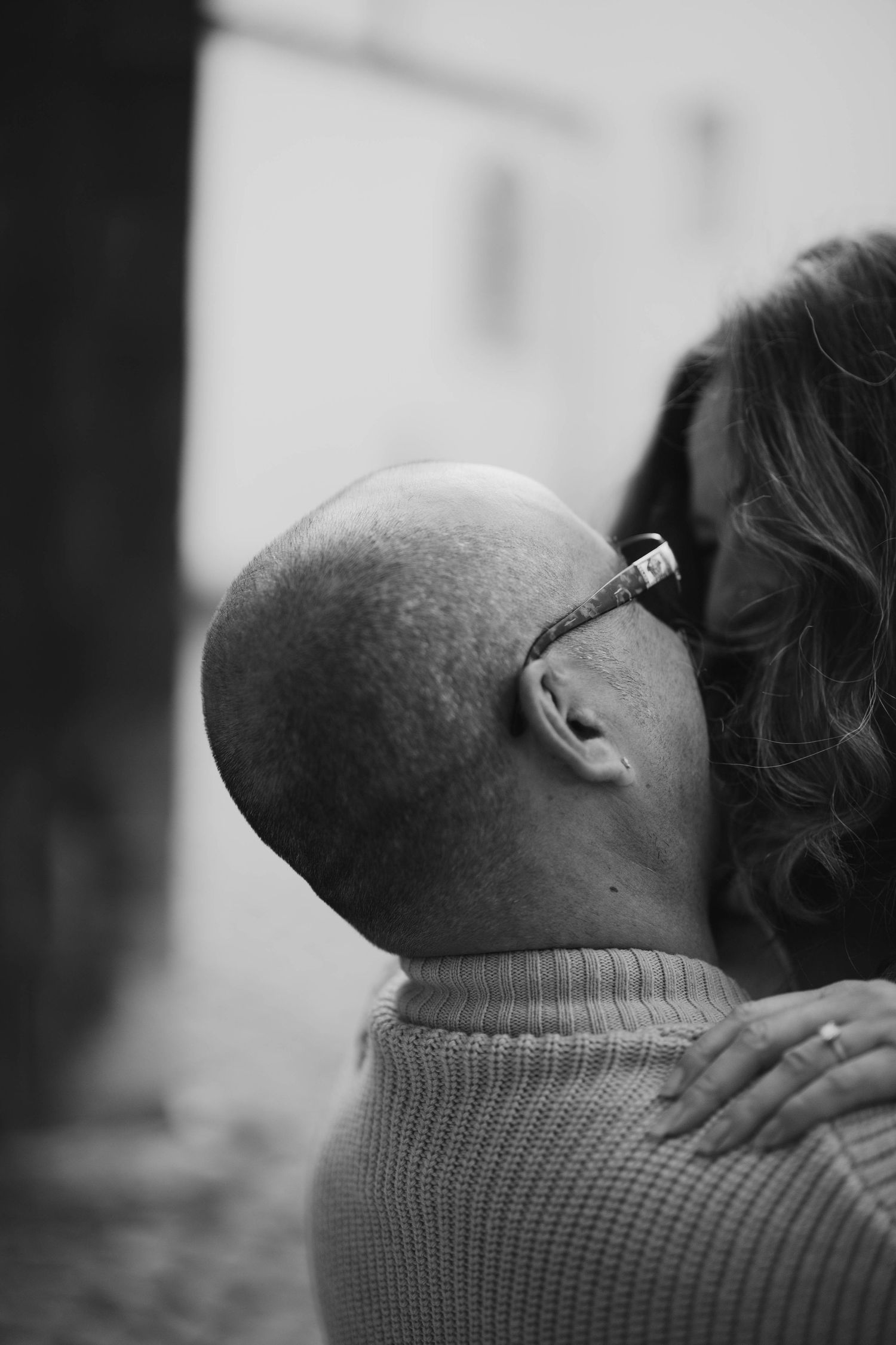 Black and white photo of a bald man with glasses embracing a woman, viewed from behind.