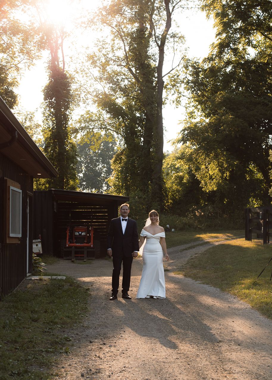 Bride and groom holding hands on a sunlit gravel path surrounded by lush trees and a rustic red barn.