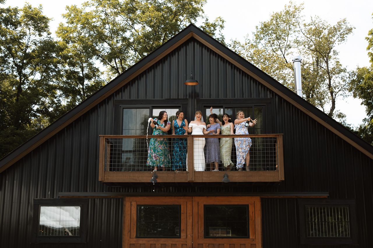 Group of women posing on a wooden balcony of a dark barn-style cabin surrounded by lush green trees.