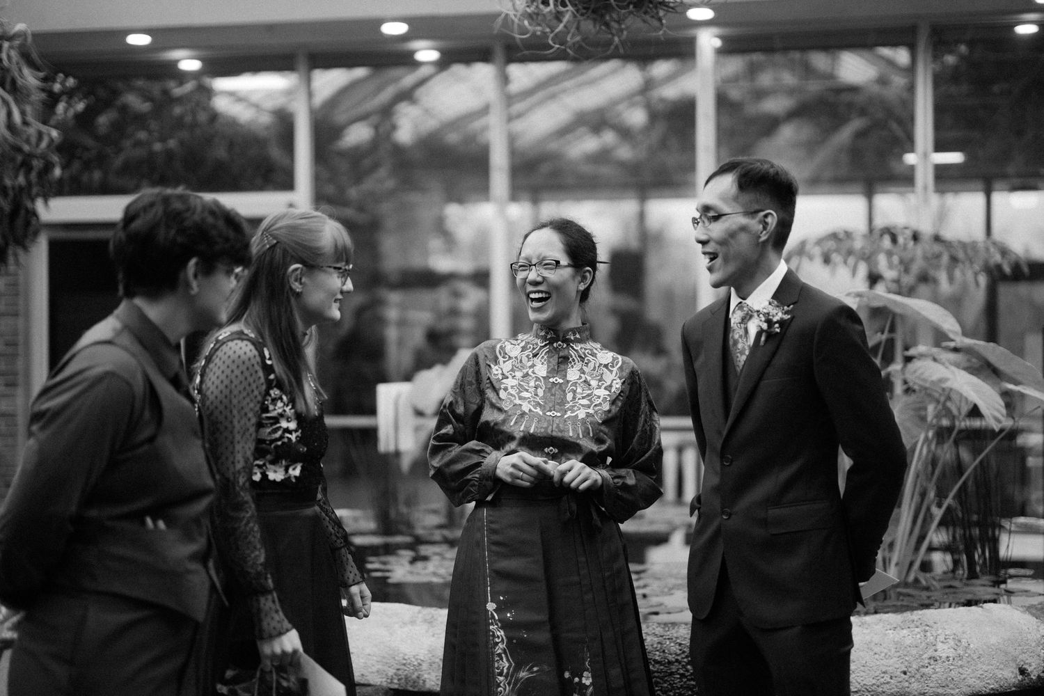 Four people laugh together outdoors in black and white photo, woman in traditional dress with suited guests.