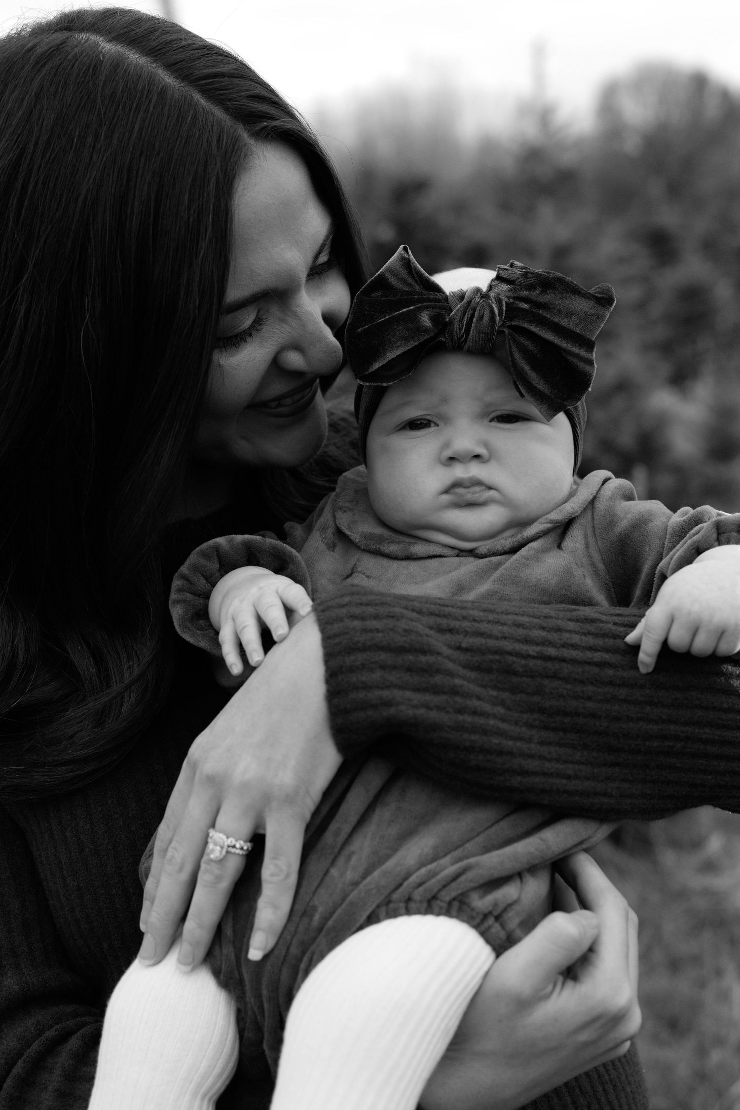 Black and white photo of a smiling mother holding her chubby baby girl wearing a large bow headband outdoors.
