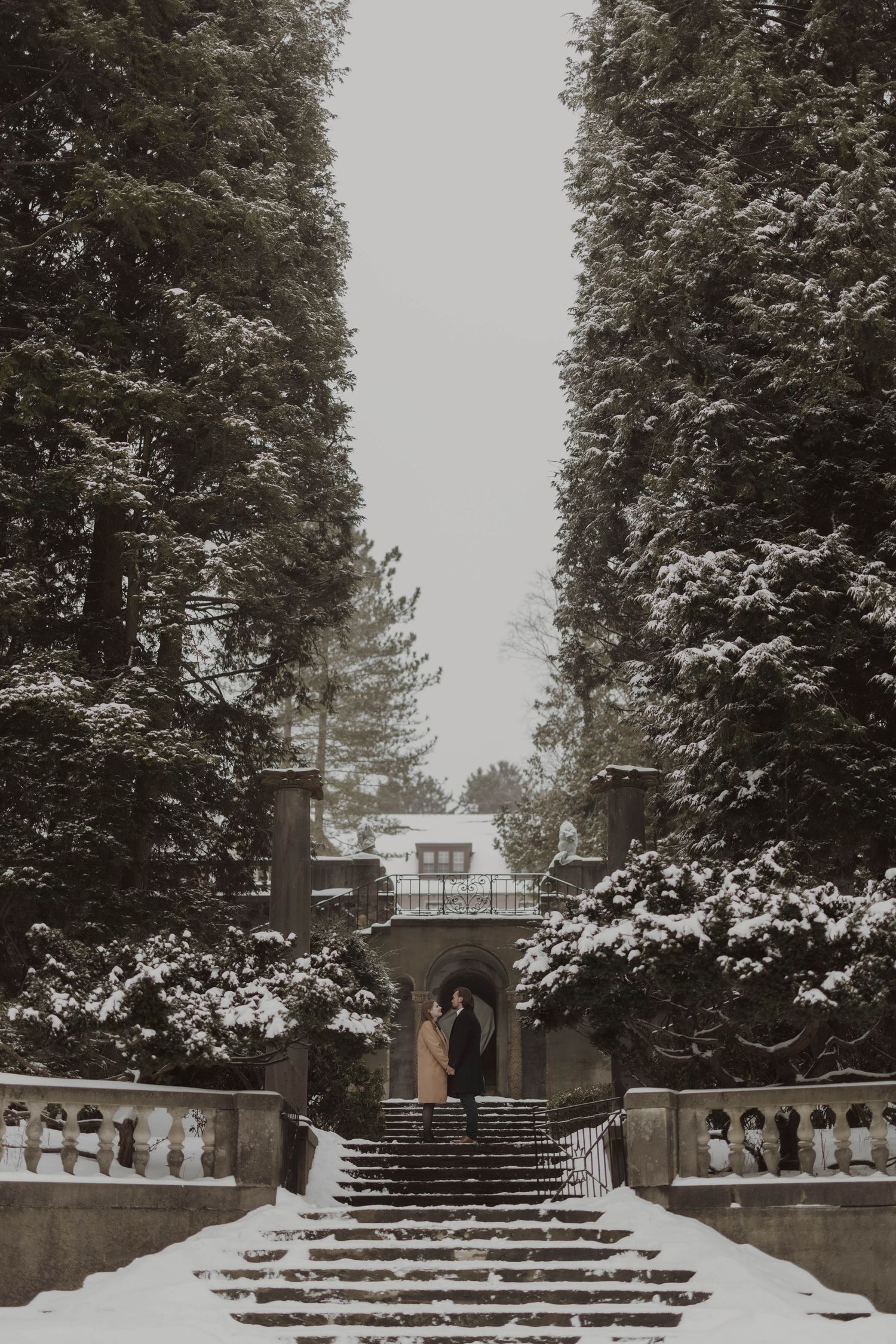 Couple embracing on snow-covered stone steps flanked by tall evergreen trees leading to a historic mansion in winter.