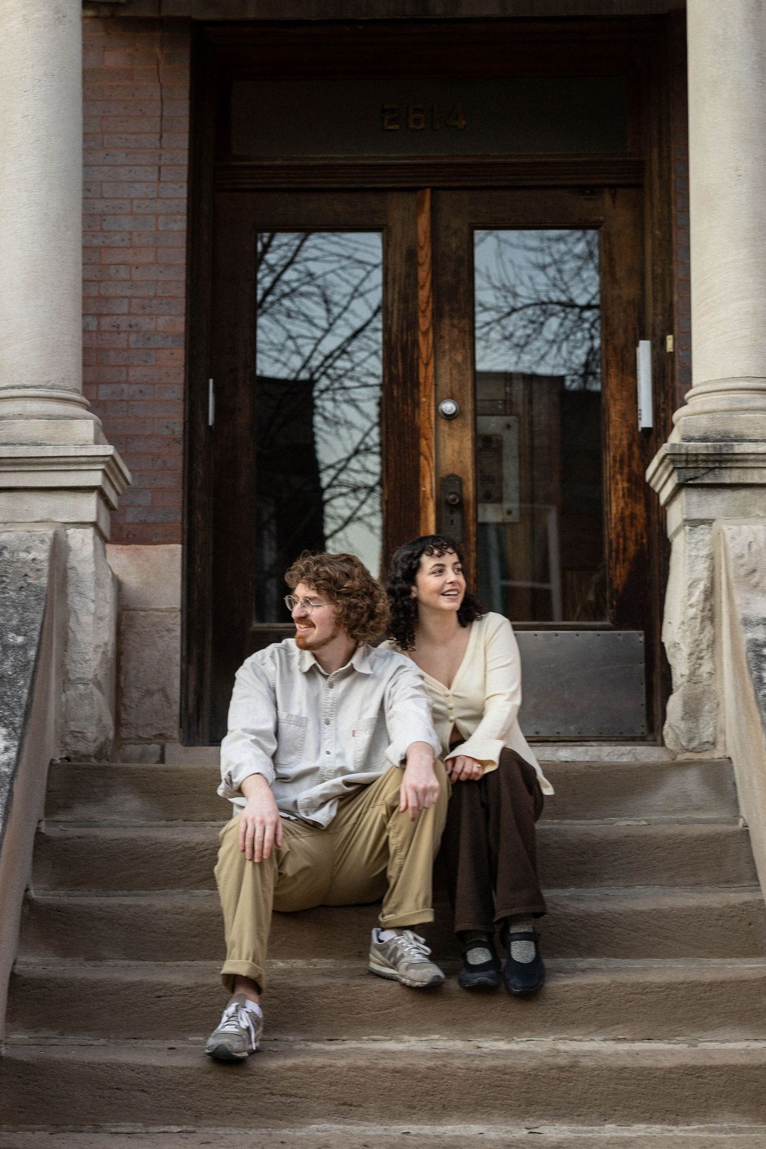 A couple sits together on stone steps outside a brick building with wooden doors, smiling warmly on a cool day.