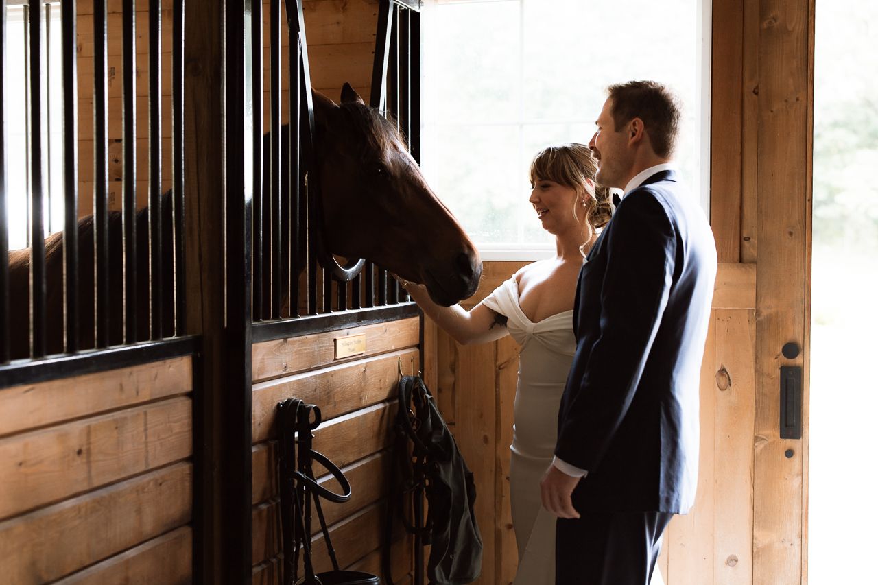 A bride and groom in wedding attire lovingly pet a dark horse in a rustic wooden stable.