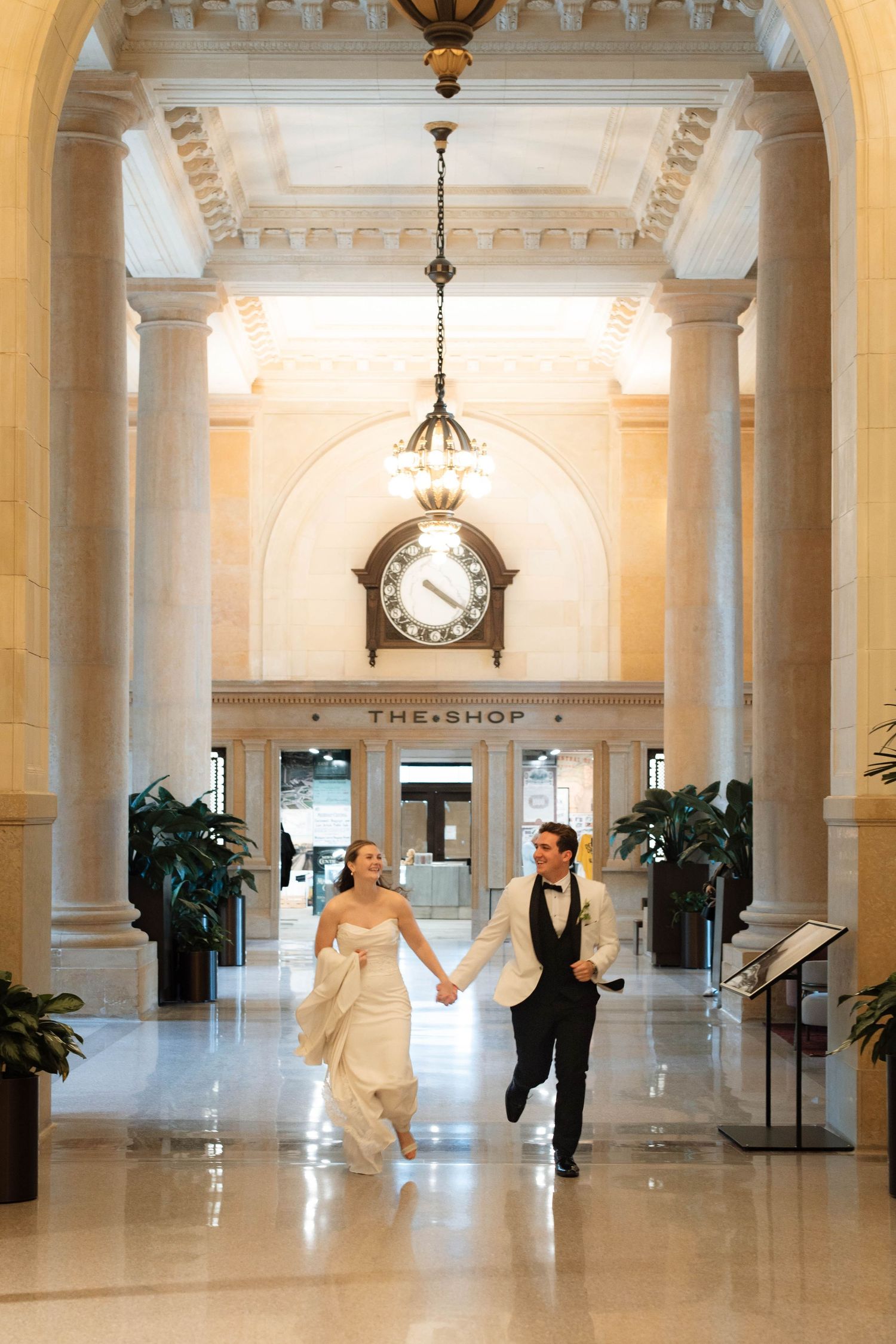 Bride and groom hold hands running through elegant marble hall with arched columns and vintage clock.