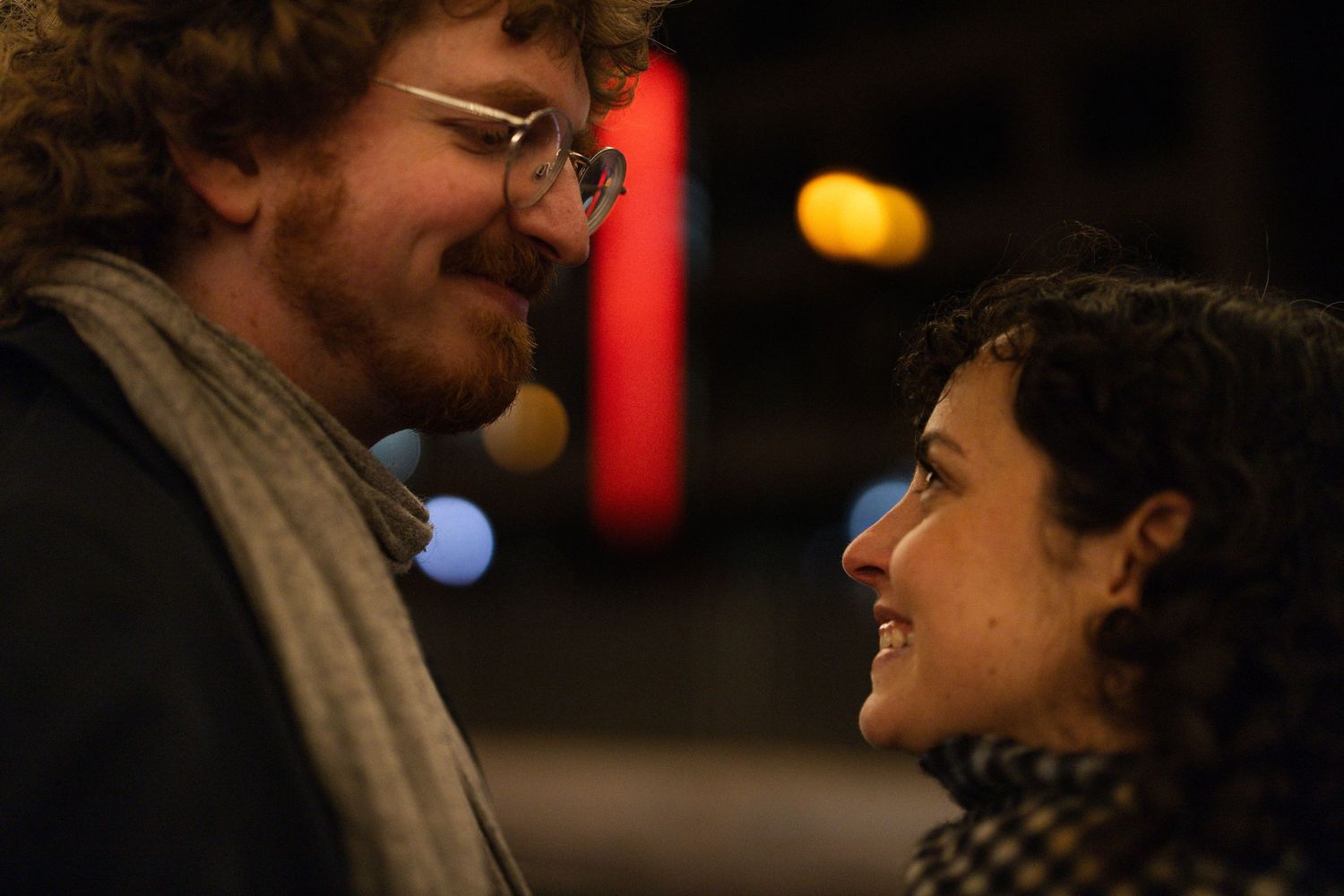 A man and woman share an intimate moment, smiling at each other outdoors at night with colorful bokeh lights behind them.