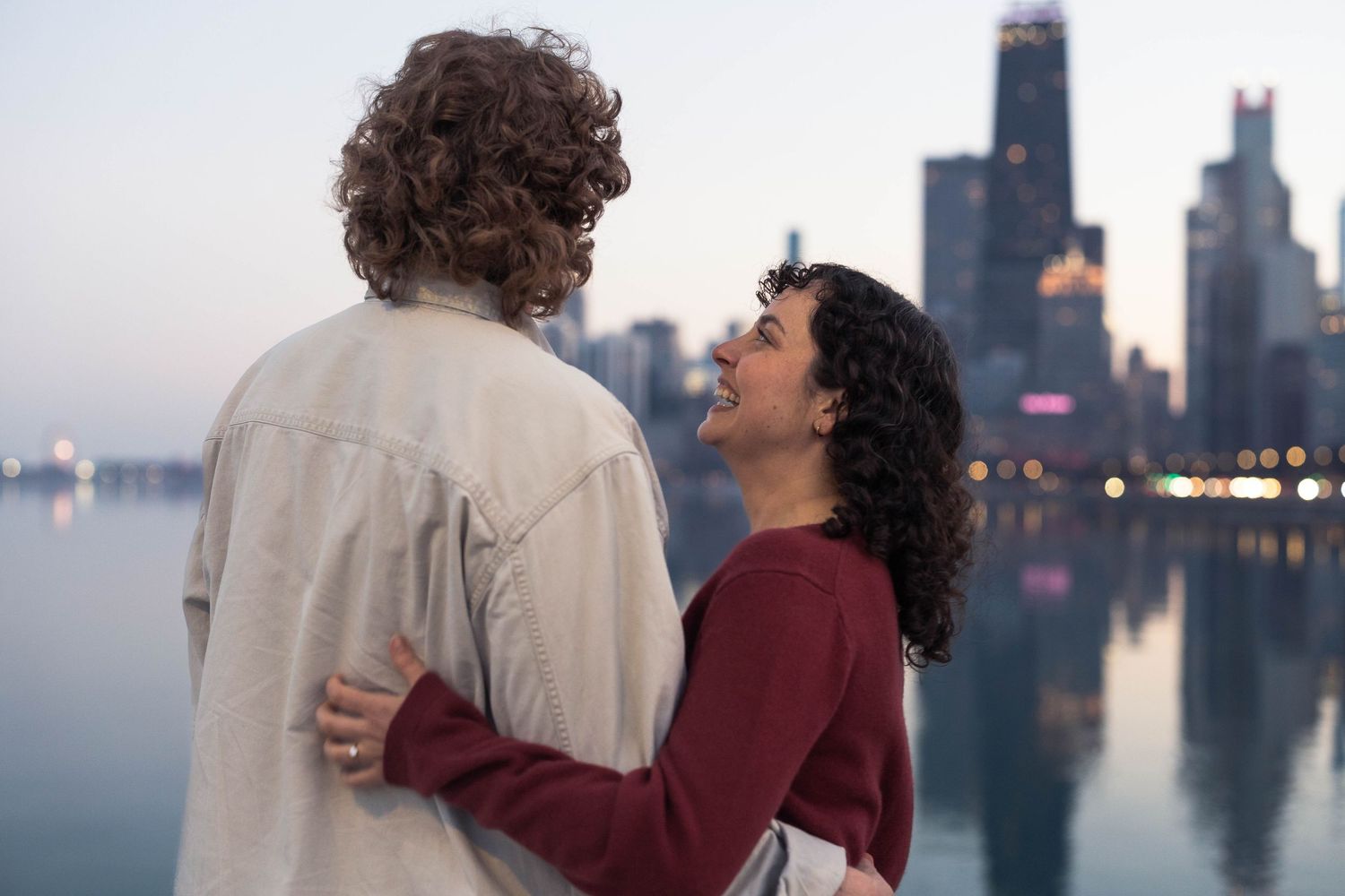 A couple embraces and smiles while standing by a city waterfront at dusk with a glowing skyline in the background.