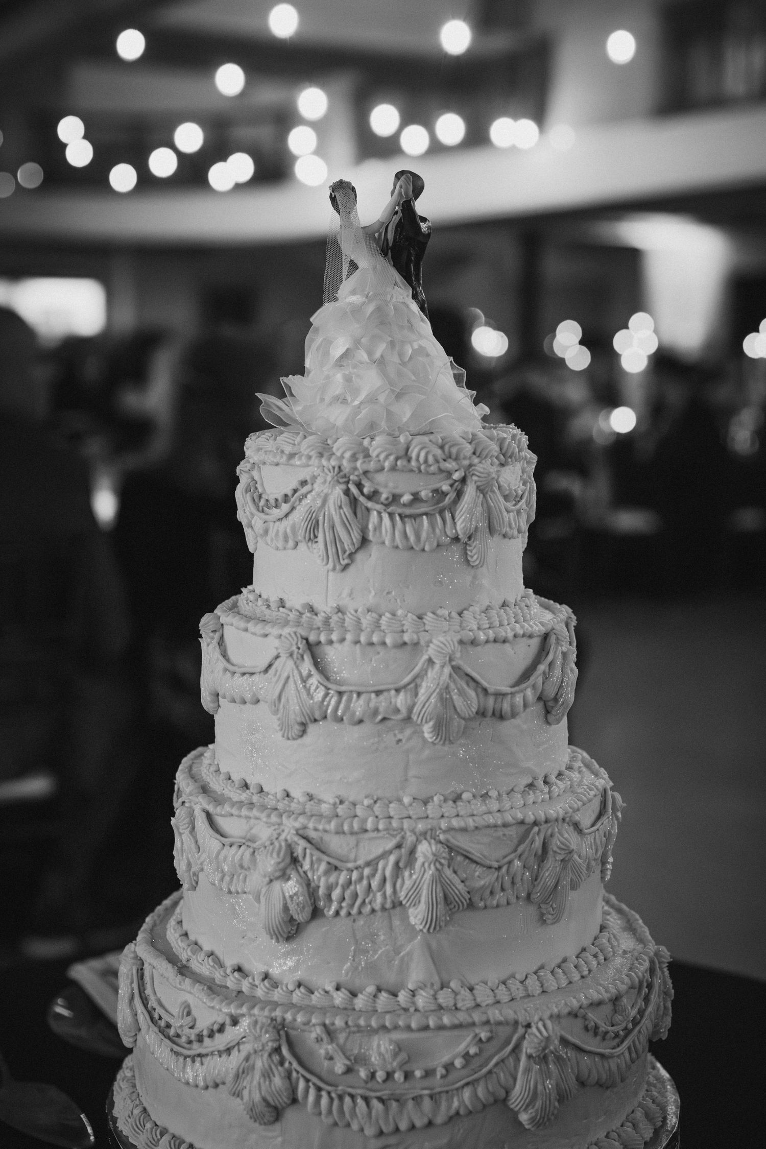 Classic four-tier white wedding cake topped with bride and groom figurines and bokeh lights.
