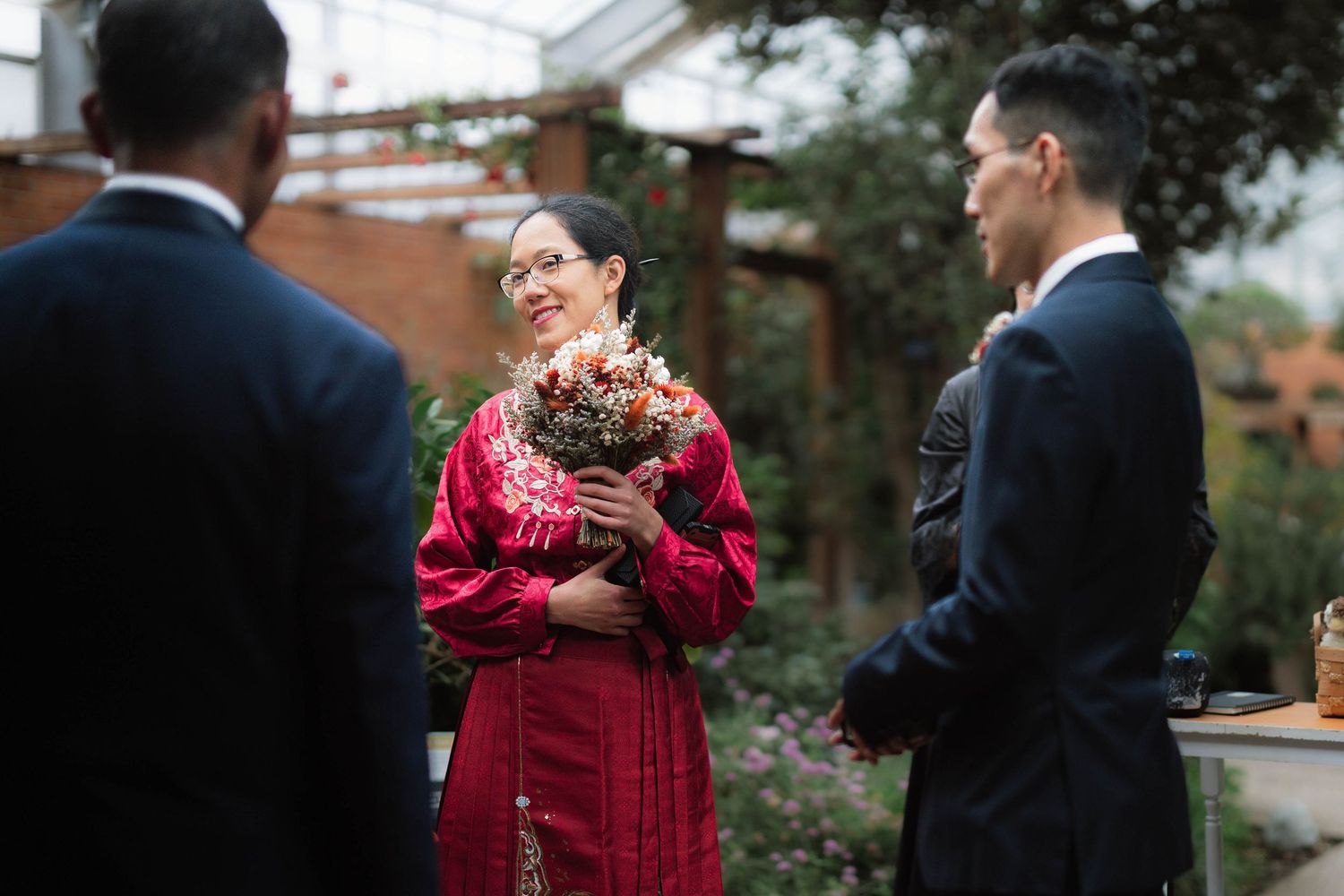 Woman in red dress holding bouquet smiles at man in suit during outdoor wedding ceremony.