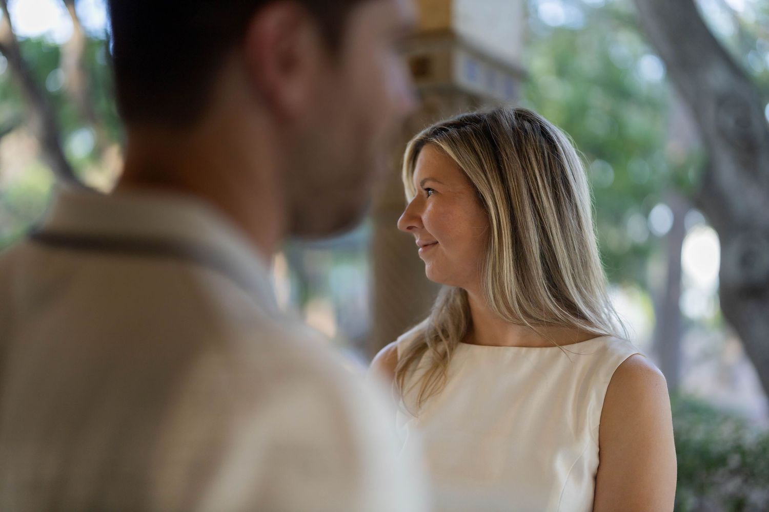 A blonde woman in a white sleeveless top gazes at a blurred man in an outdoor setting.