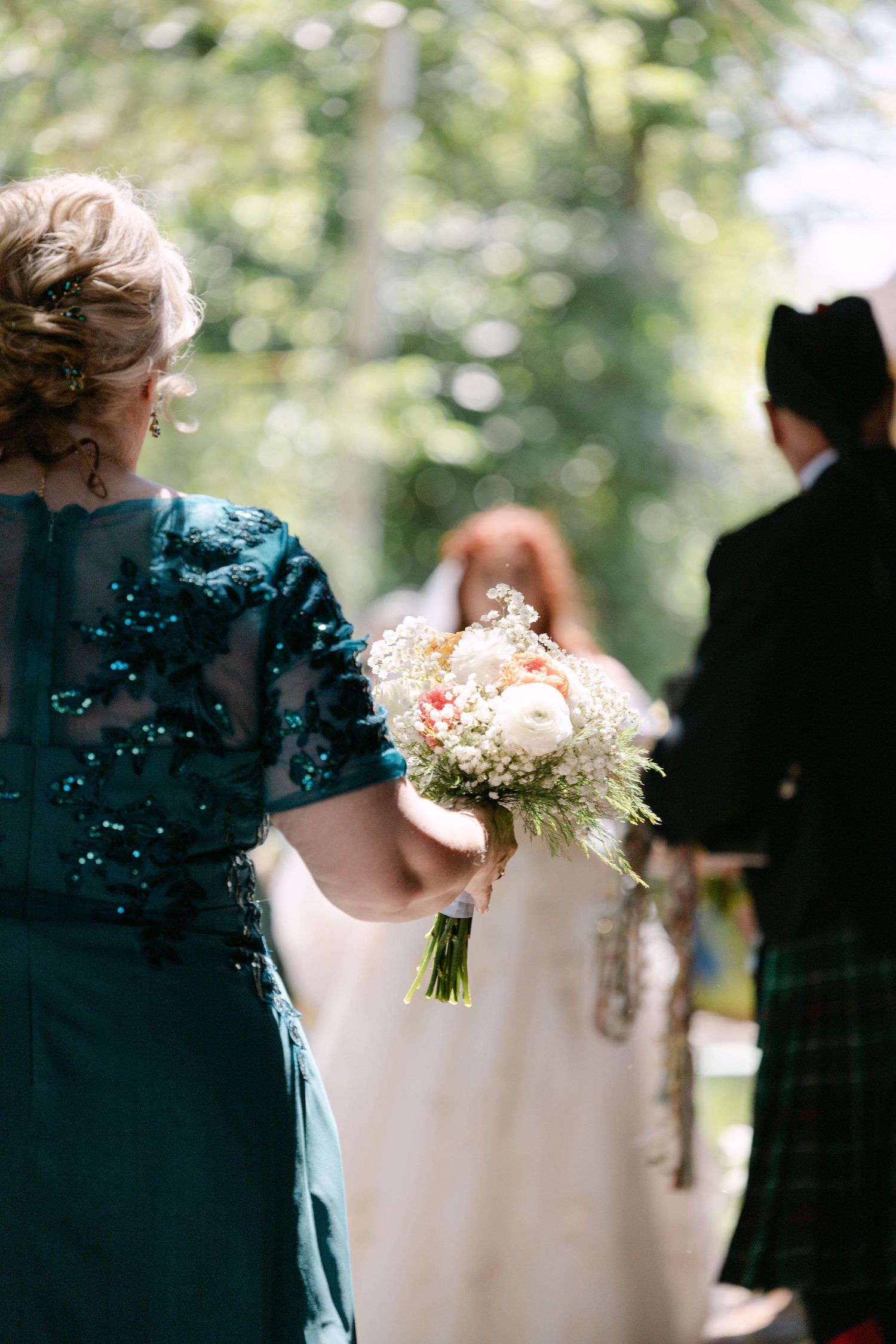 A wedding guest in a teal sequined dress holds a white floral bouquet during an outdoor ceremony.