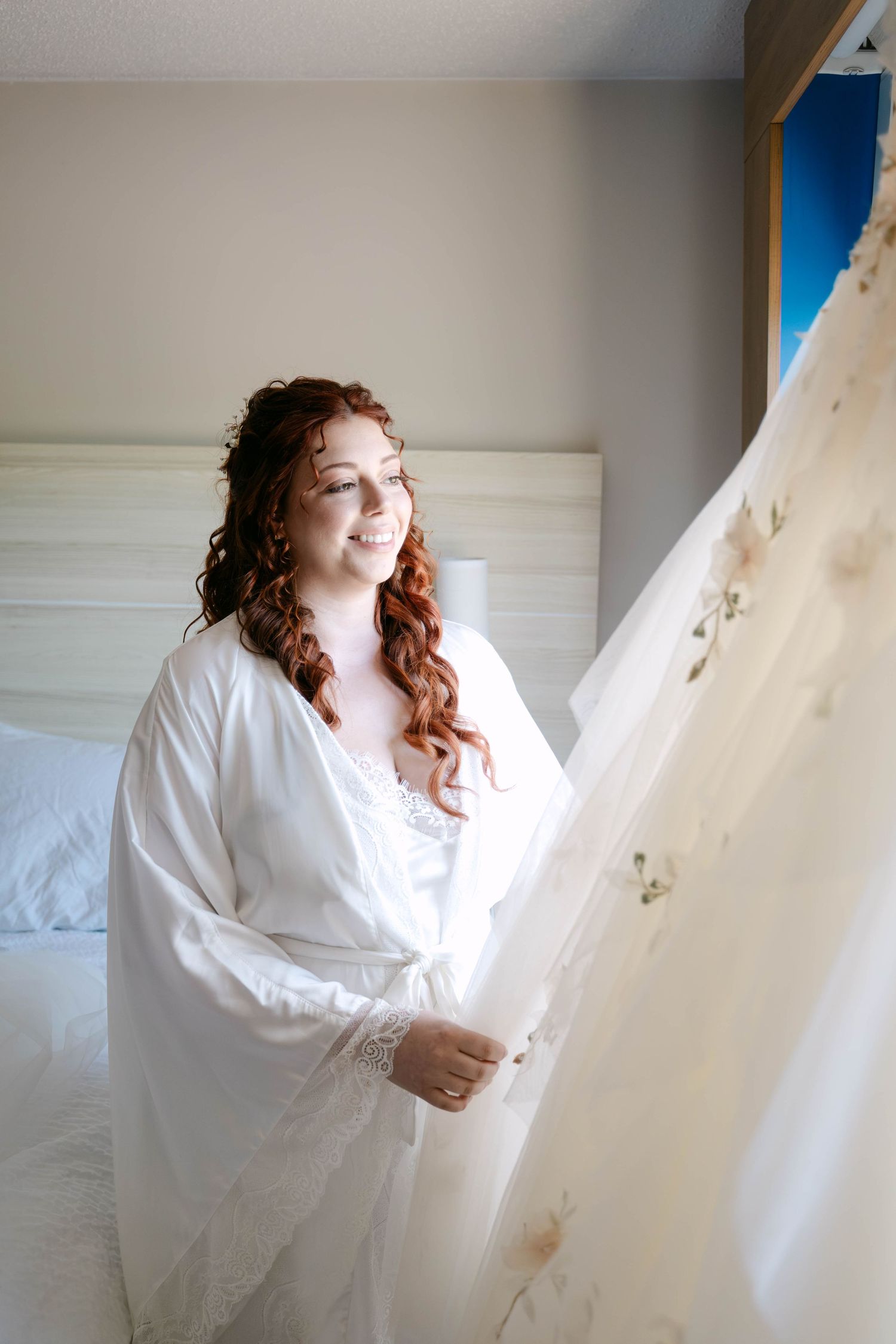 Bride in white robe admiring her wedding dress in a bright bedroom before the ceremony.
