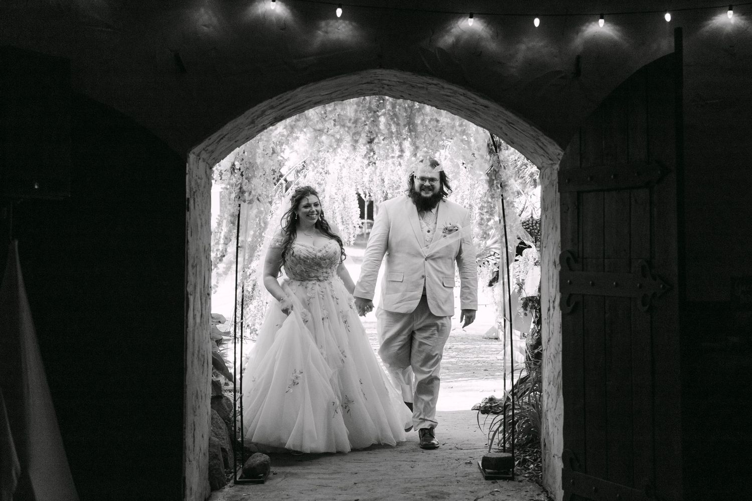 Black and white photo of a bride and groom holding hands walking through a stone archway at their wedding.
